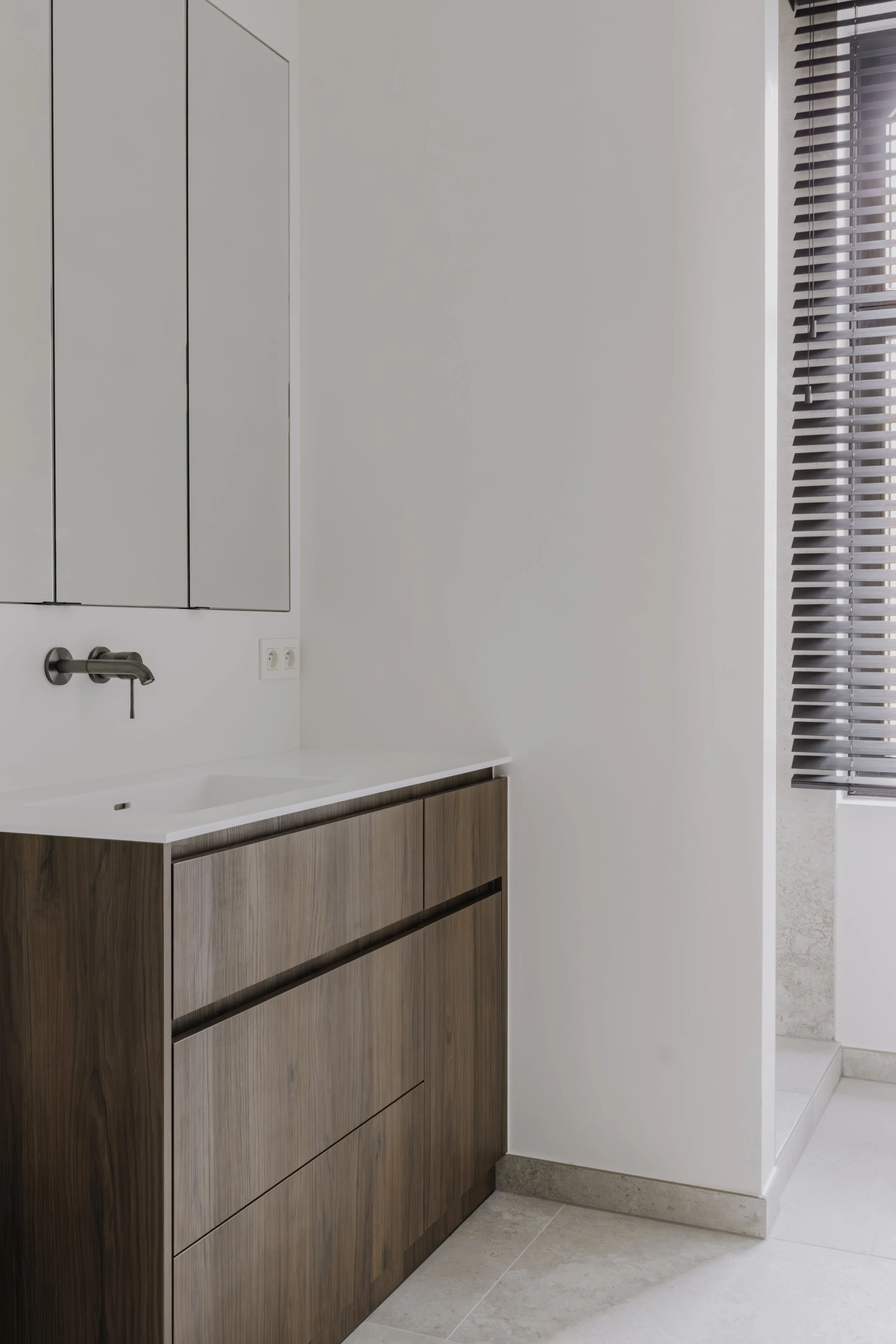 Moderne badkamer met wit werkblad, houten ladekast, wandkraan en natuurlijk licht.