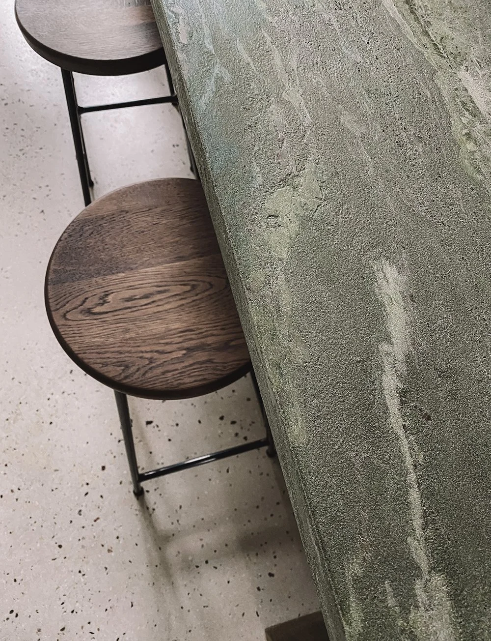 Interieurdetail van een groen natuurstenen werkblad met houten barkrukken en gespikkelde vloer.