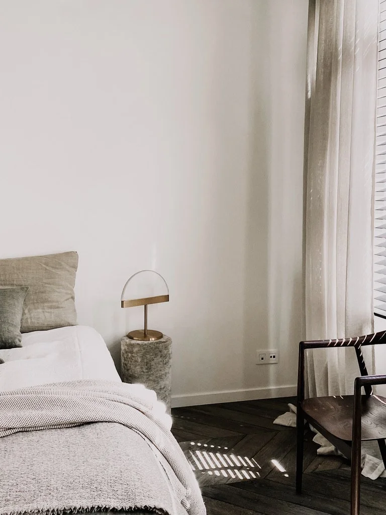 Sfeervolle slaapkamer met bed, houten stoel, moderne bijzettafel en zacht daglicht door transparante gordijnen.
