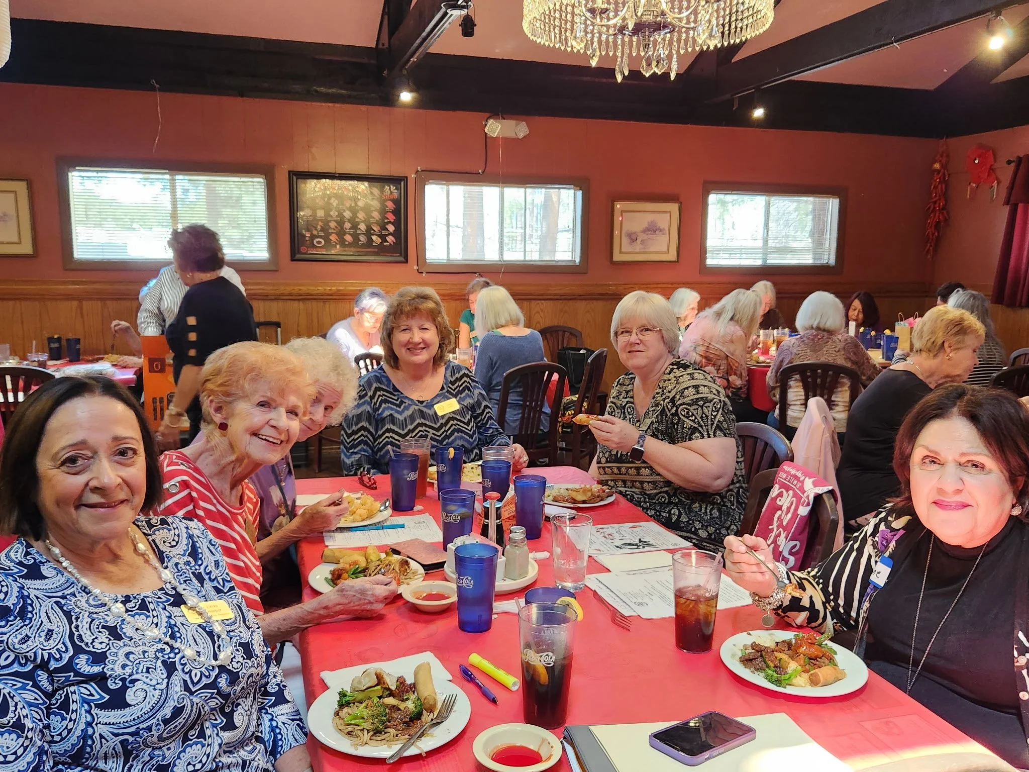 A group of elderly women sitting around a banquet table enjoying a meal, with plates of food, drinks, and festive decorations in a restaurant or banquet hall.