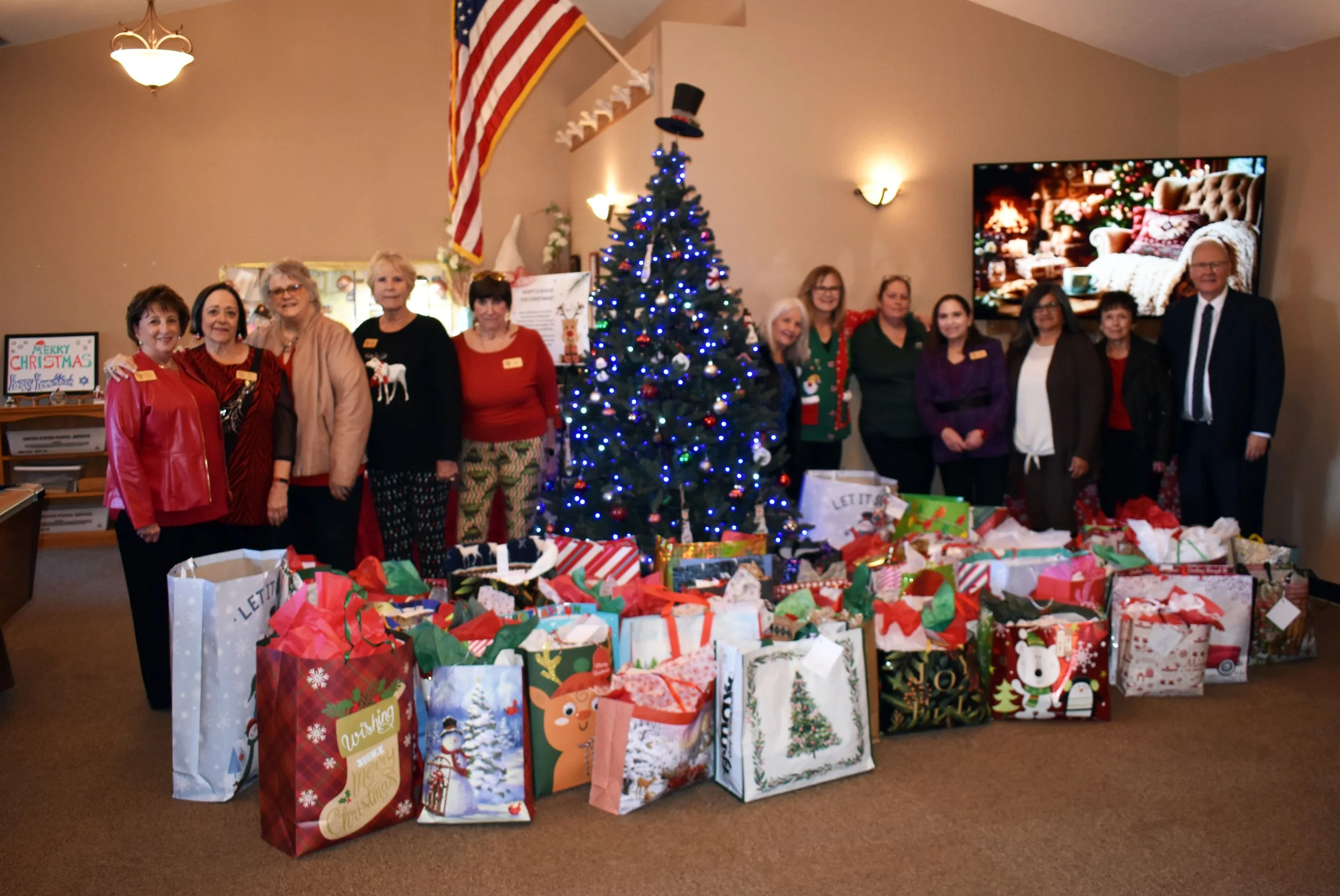Group of people standing in front of a decorated Christmas tree with wrapped presents below, in a room with beige walls and a television displaying a cozy fireplace scene.