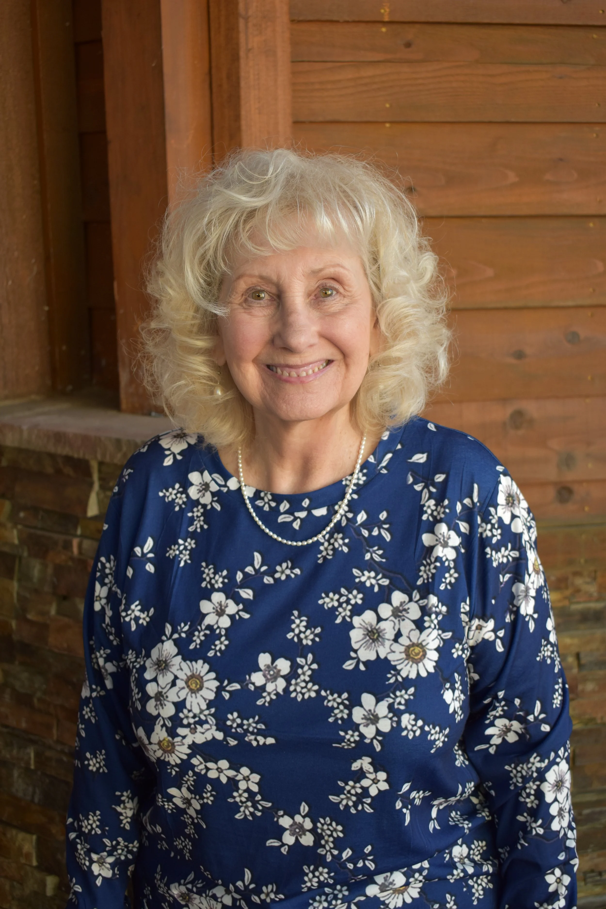 A smiling senior woman with curly gray hair wearing a blue dress with white floral pattern and a pearl necklace, standing in front of a wooden wall.