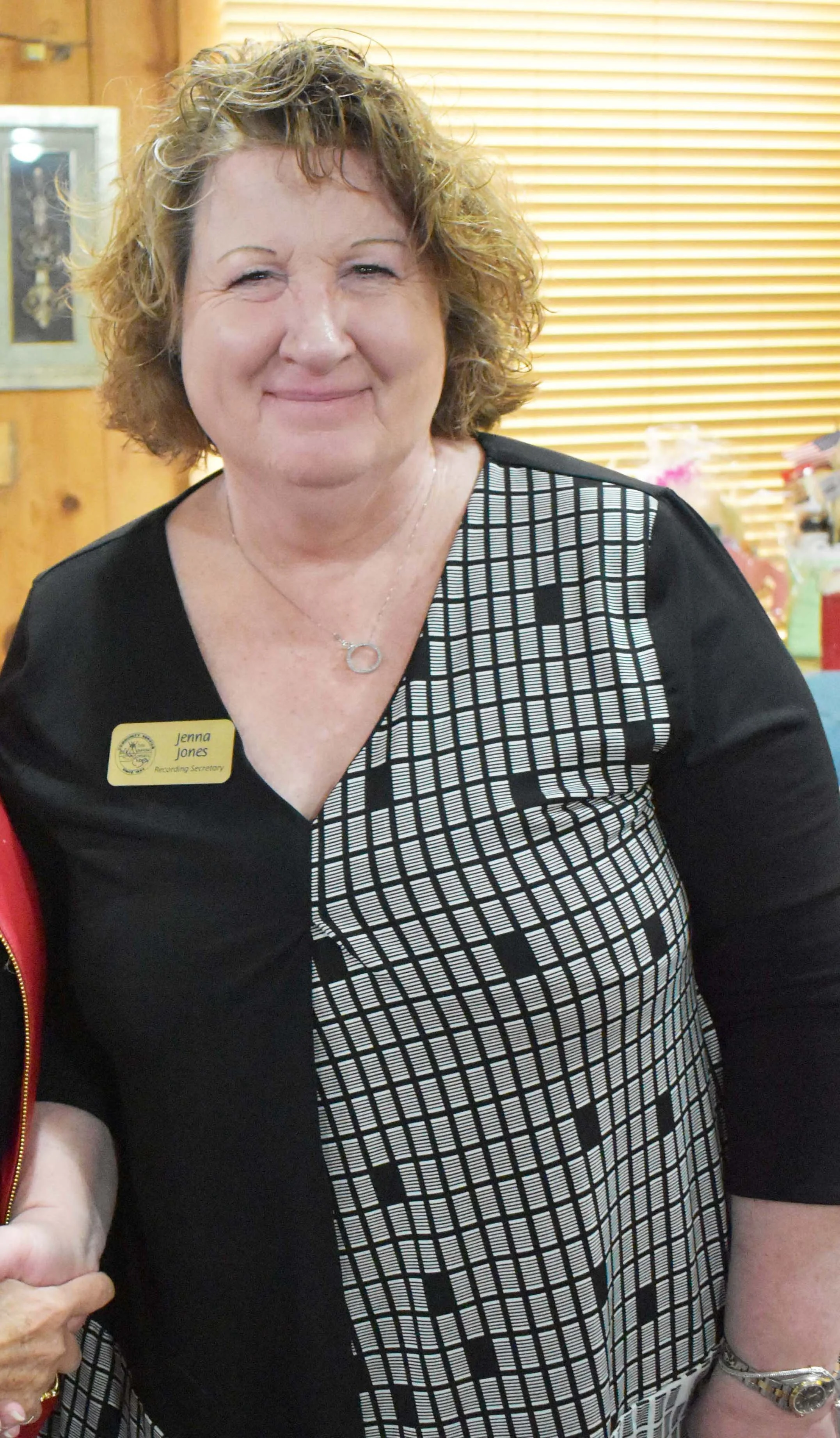A smiling woman with curly short hair wearing a black and white checked dress, a name tag, and a necklace, standing indoors with wooden walls and window blinds in the background.