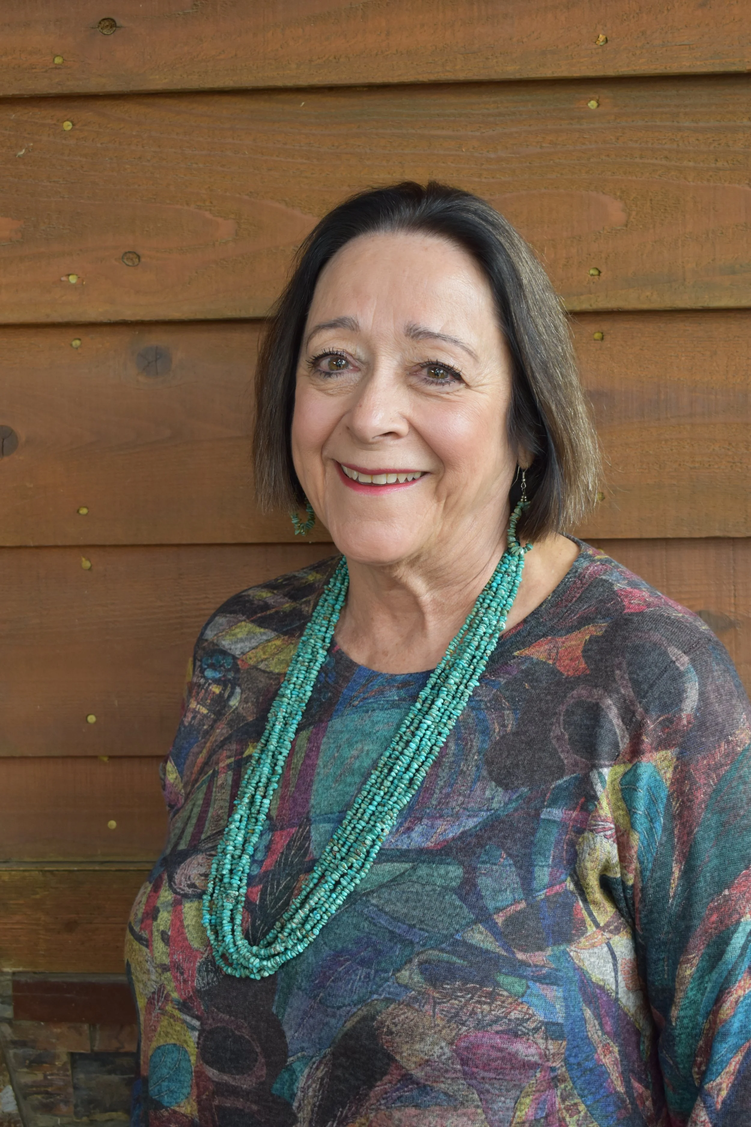 A woman with short dark hair, wearing a colorful patterned top and multiple turquoise necklaces, smiling in front of a wooden wall.