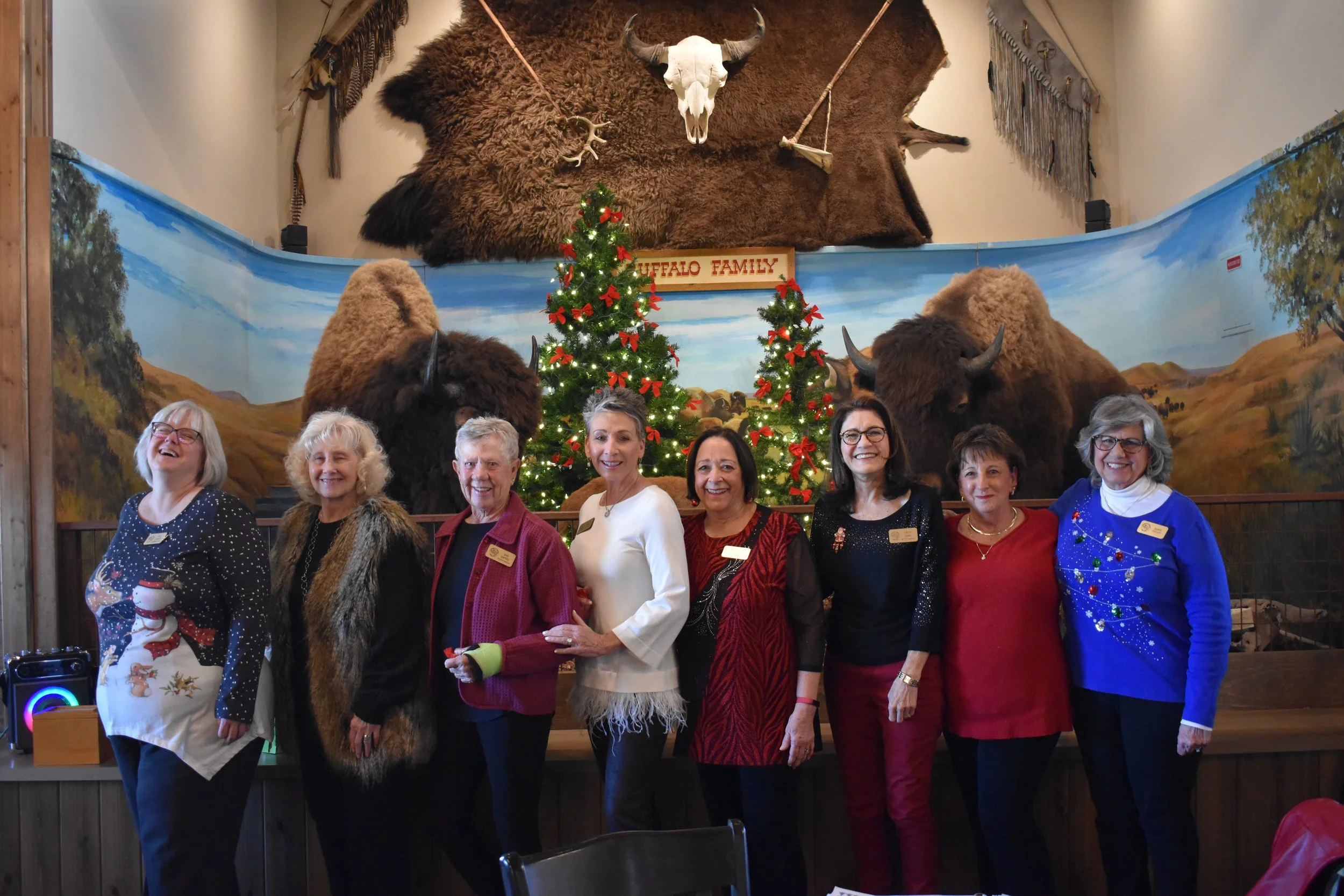 A group of nine women standing in front of Christmas decorations including a small decorated Christmas tree, festive sweaters, and holiday accessories, with a painted landscape mural and mounted animal skulls and fur above them.