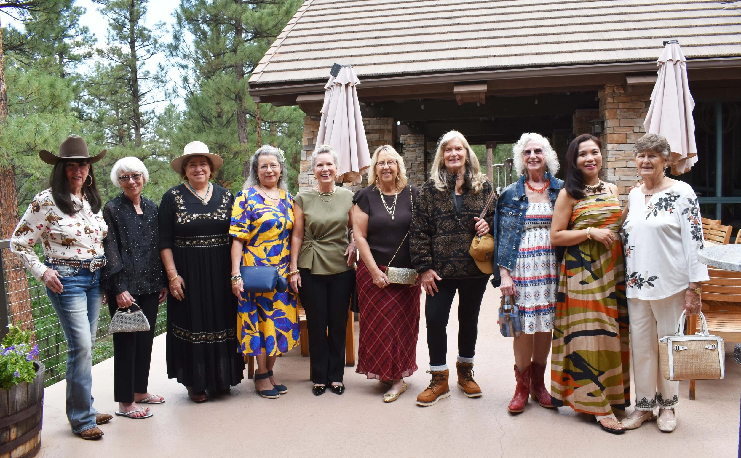 Group of women standing together outdoors, dressed in colorful and stylish outfits, in front of a building with a stone facade and surrounded by trees and greenery.
