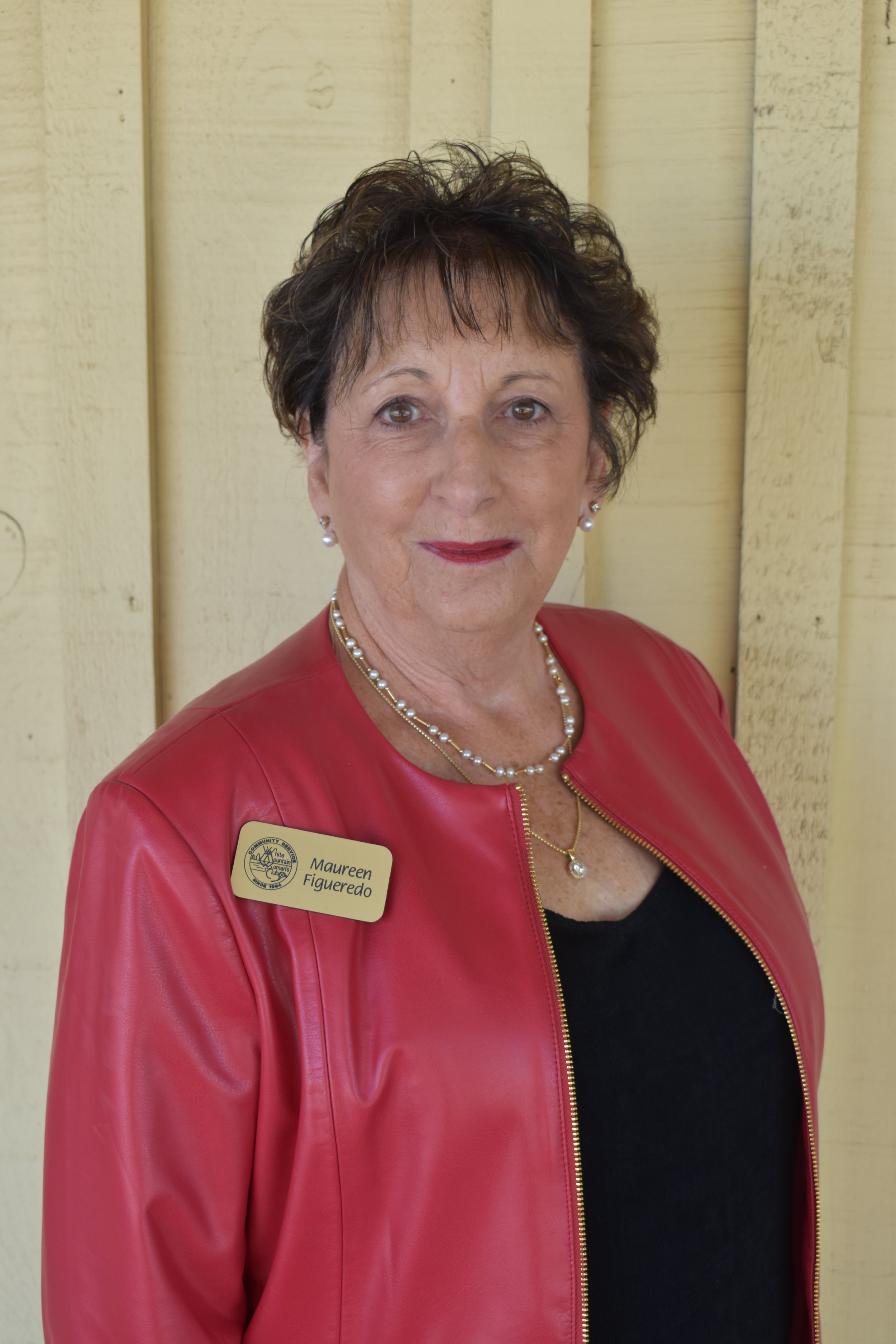 Close-up of a woman wearing a red leather jacket, pearl necklace, and pearl earrings, standing against a light wooden background.