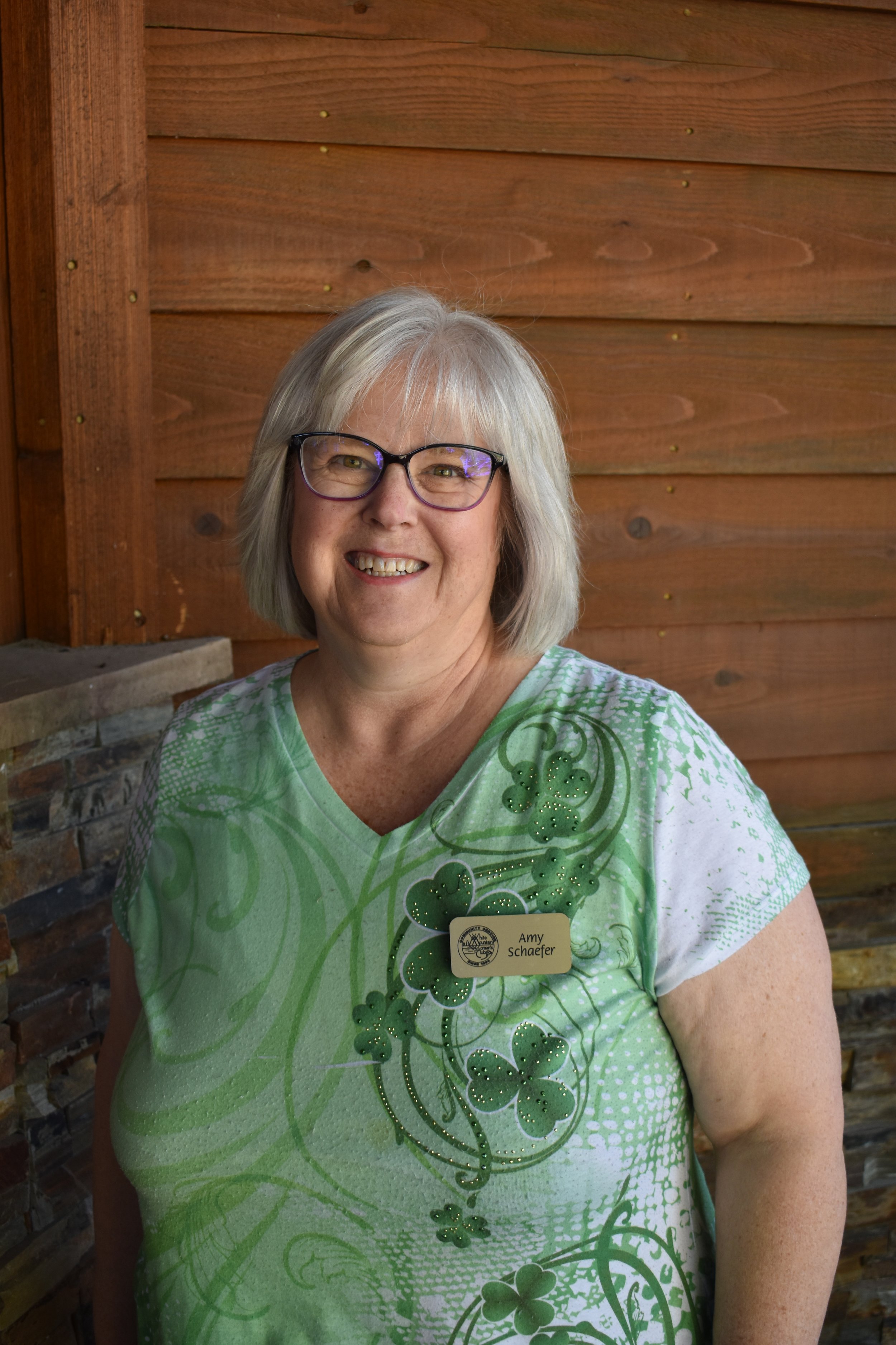 A smiling woman with gray hair and glasses, wearing a green and white shirt with a shamrock design, standing in front of a wooden wall.