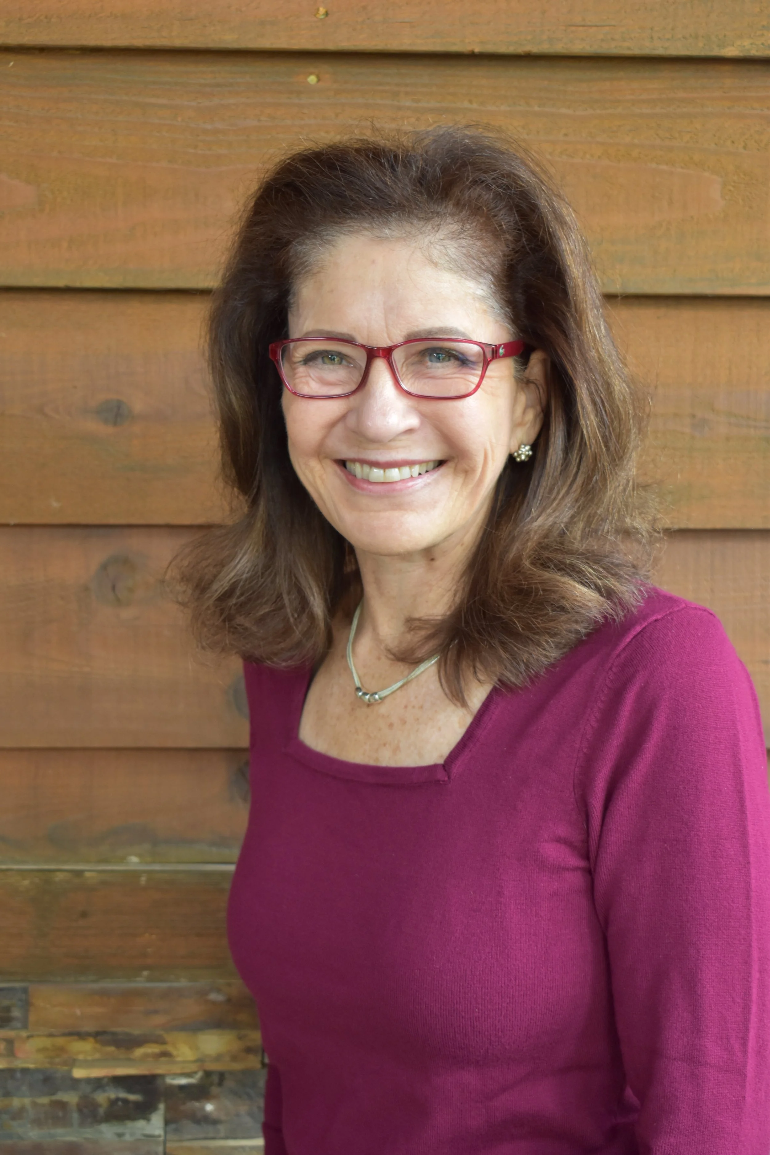 A woman with brown hair wearing red glasses, a purple top, and a silver necklace standing in front of a wooden wall.