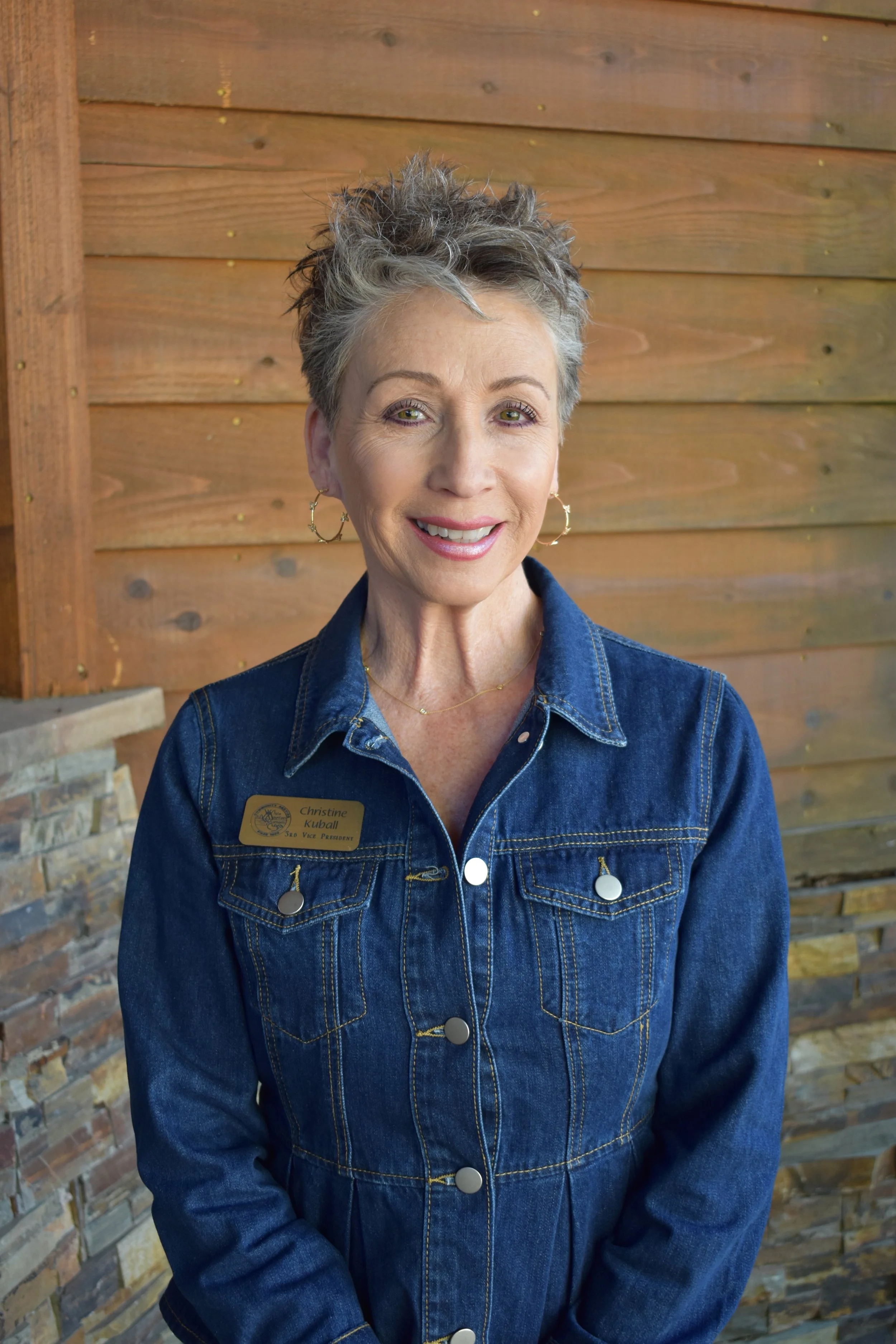 A smiling woman with short gray hair and hoop earrings wearing a denim jacket standing in front of a wooden wall.