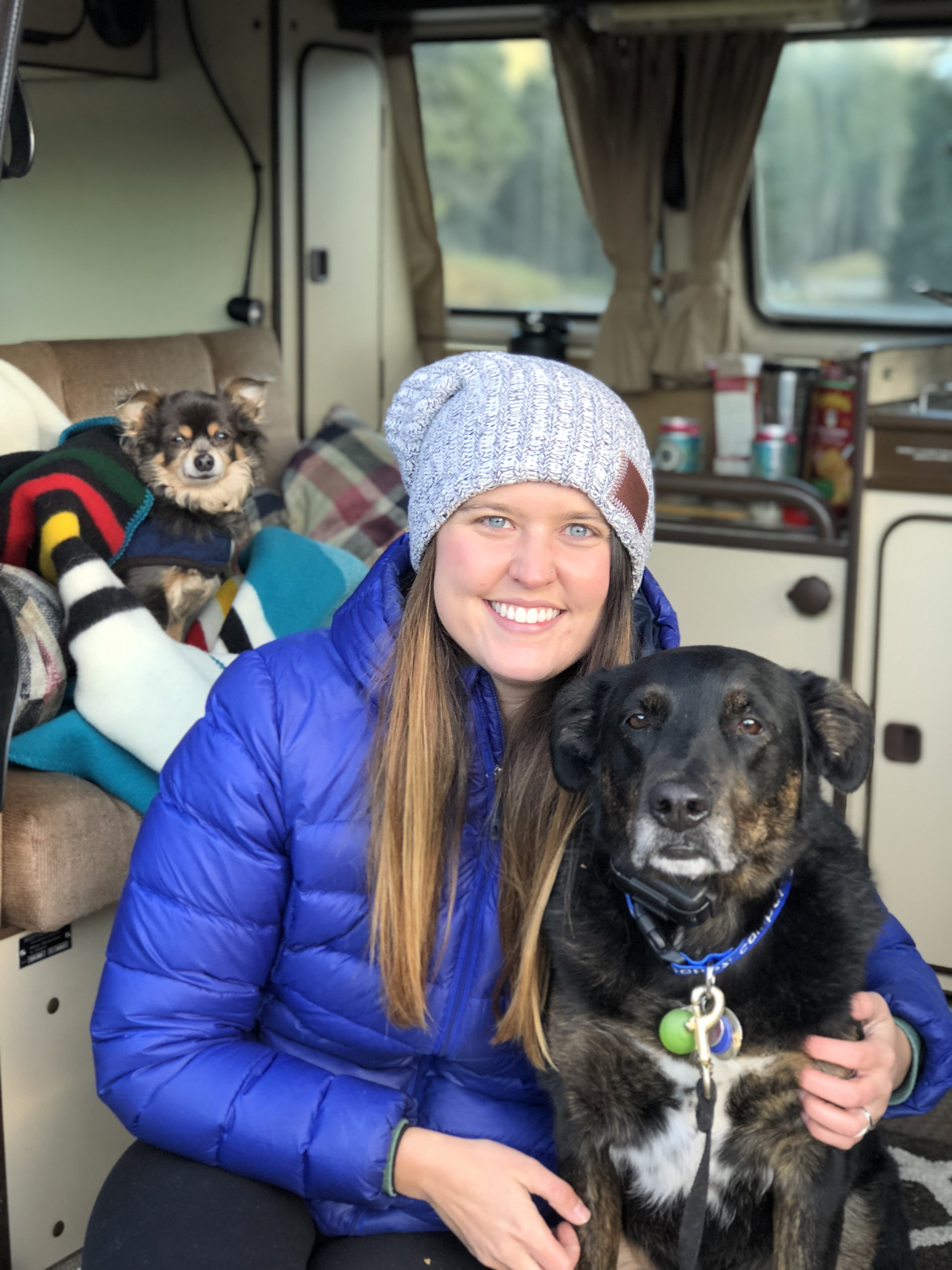 A woman with long red hair, wearing a gray knit hat and a blue puffer jacket, smiling and sitting inside a vehicle with two dogs, one on her lap and a small dog on a sofa behind her, with a blurred outdoor landscape visible through the windows.