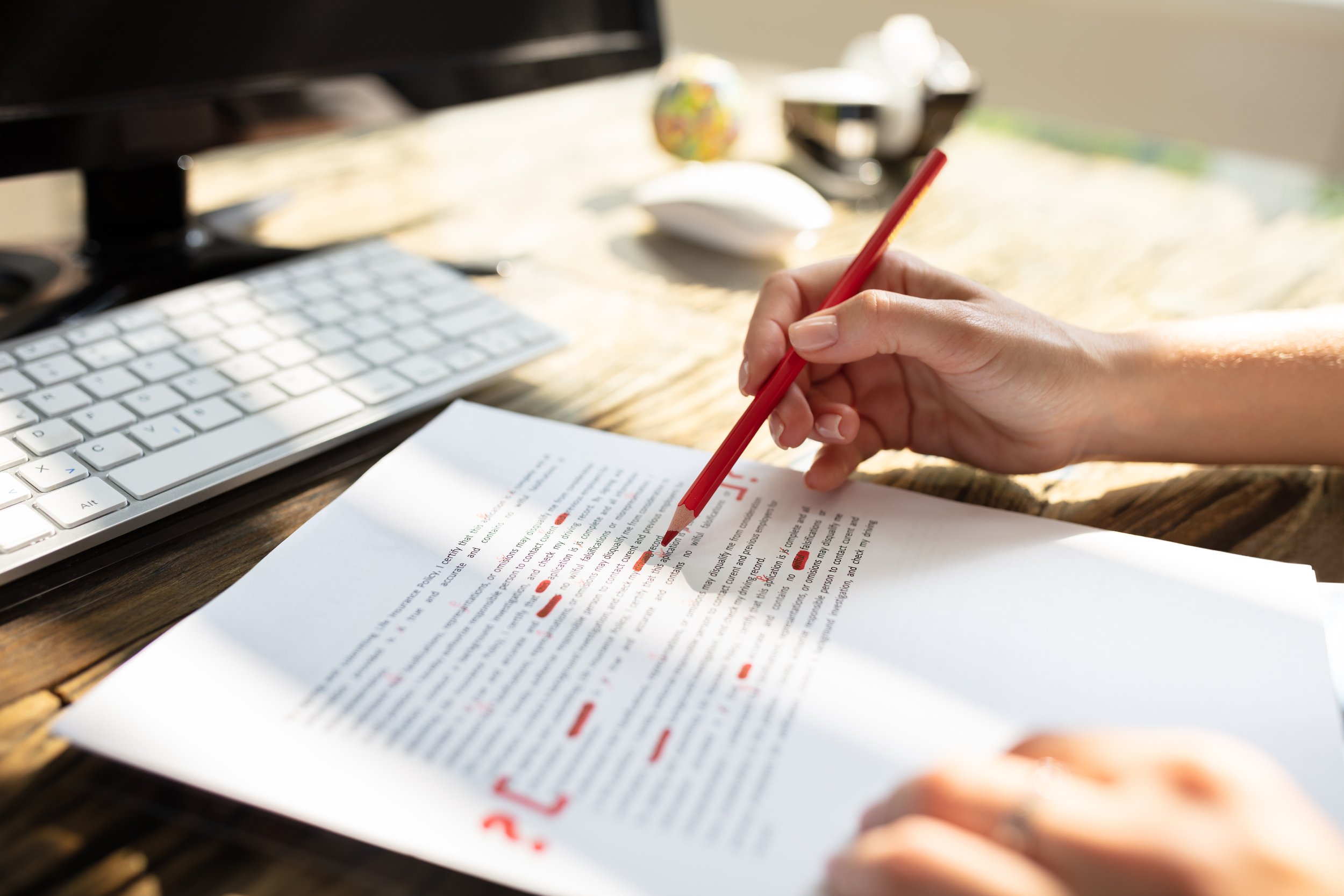 A person holding a red pen and highlighting text on a printed document with a computer keyboard and office supplies in the background.