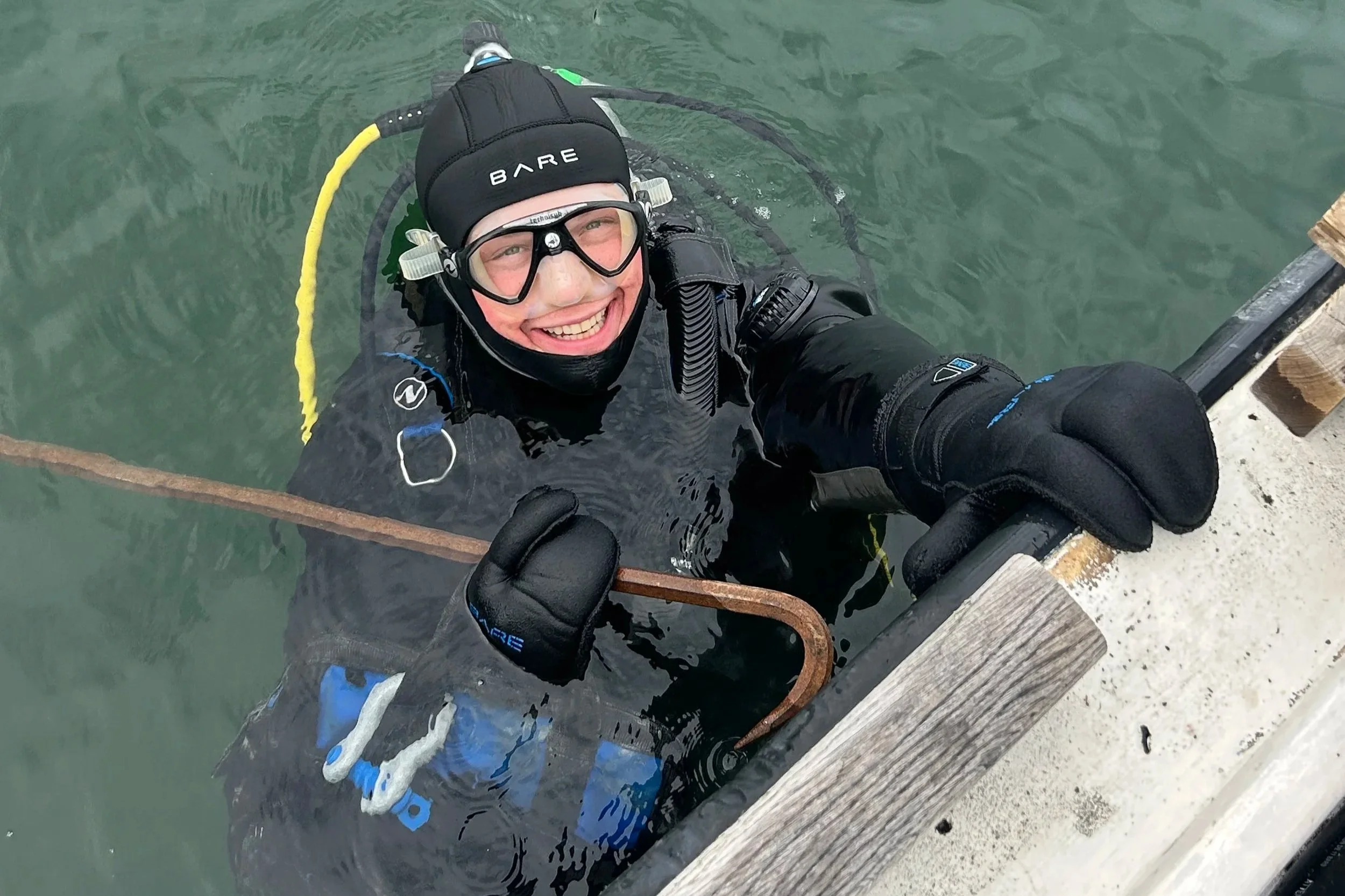 A person in scuba gear smiling while climbing out of the water onto a wooden dock.