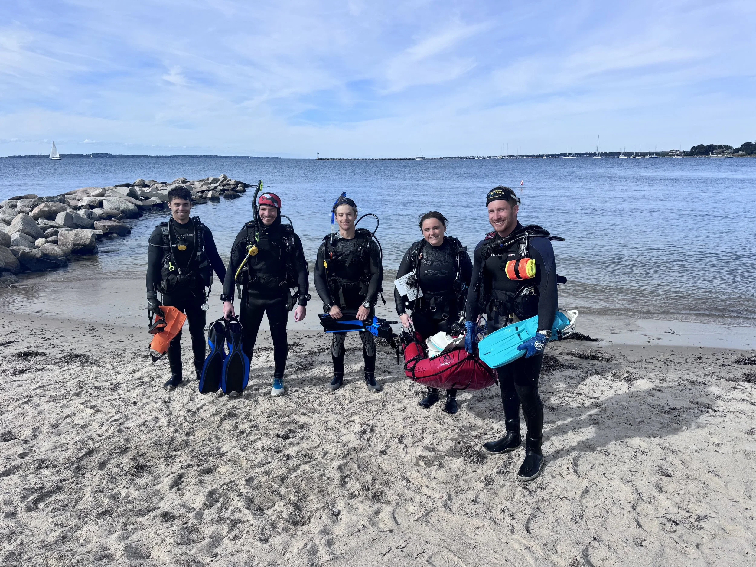 Five people standing on a sandy beach, dressed in snorkeling gear with fins and masks, smiling with a body of water and sailboats in the background.