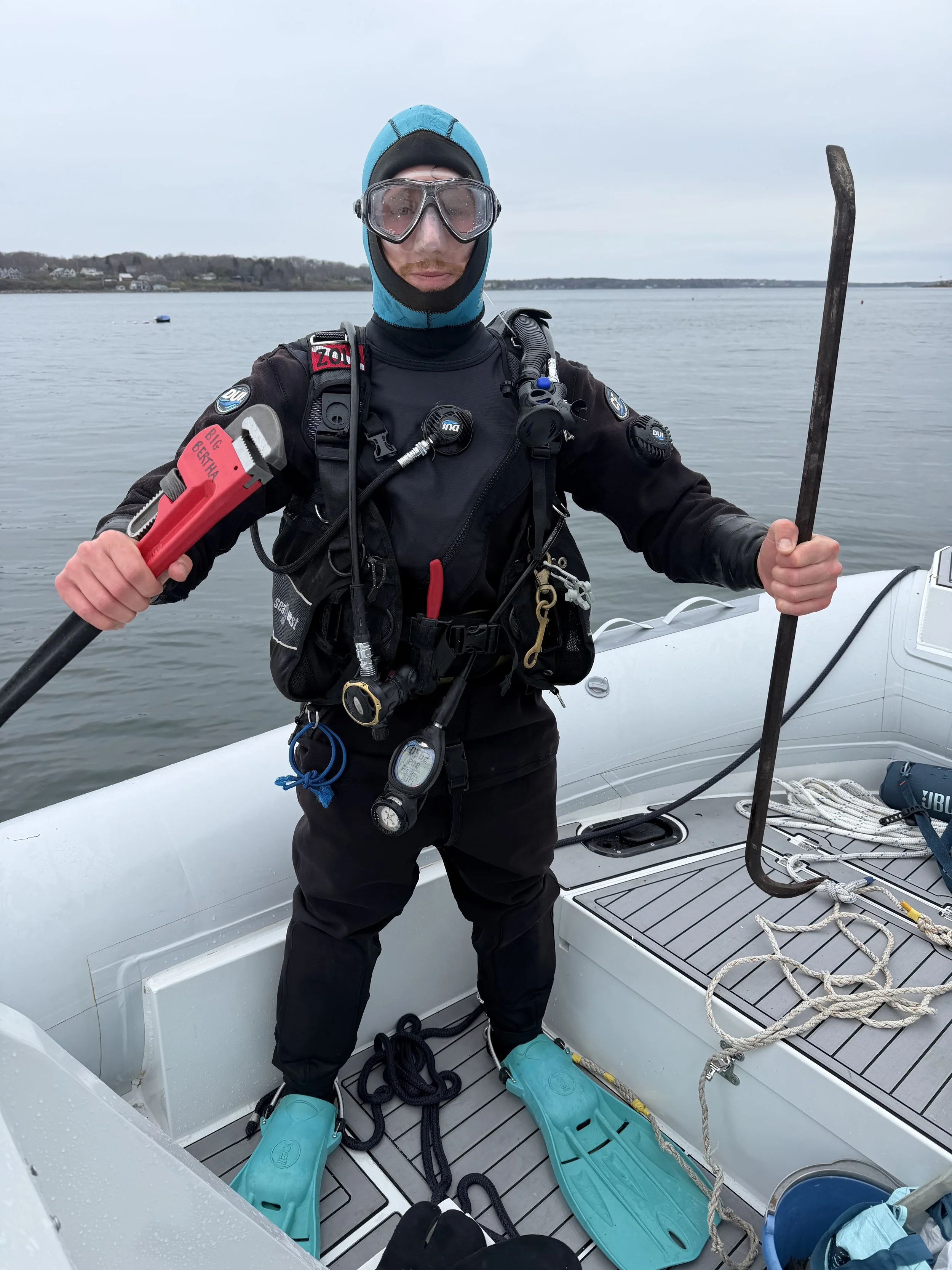 A person dressed in wetsuit with diving gear holding a metal tool and standing in a boat on water, with shoreline and houses in the background.