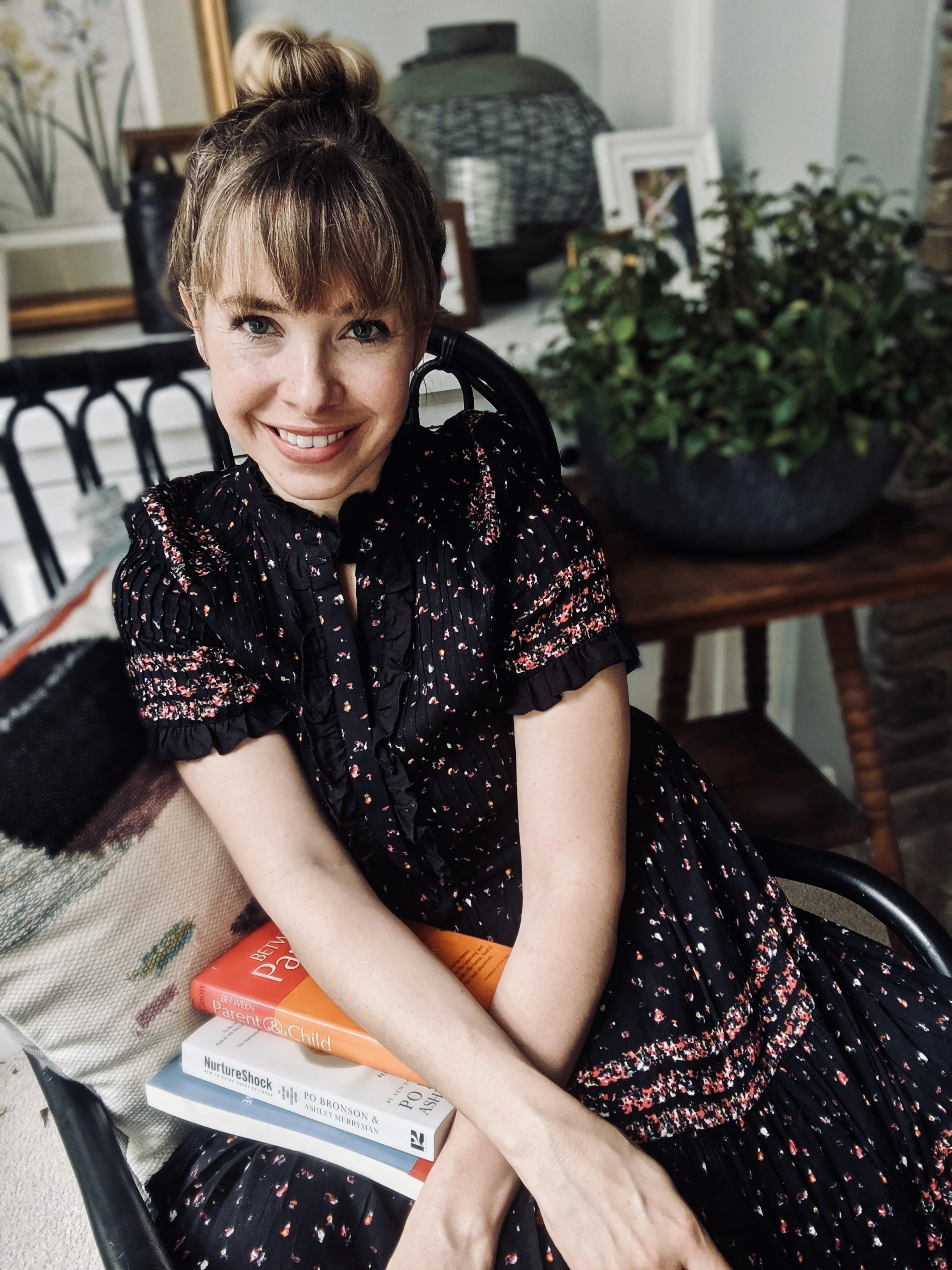 A smiling woman with reddish-brown hair in a bun sitting in a black chair, wearing a black dress with red and white floral patterns. She is holding a book titled 'Between Parent & Child,' with other books stacked underneath, and a cloth bag over her lap. There is a large potted plant behind her and various decorative items and framed pictures in the background.