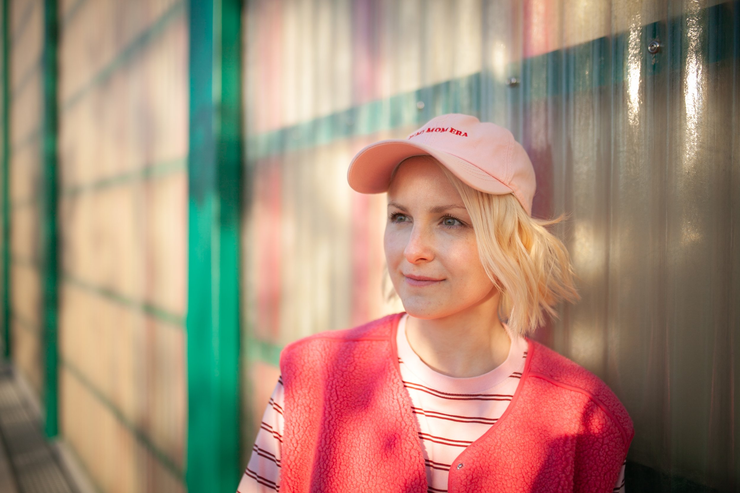 Young woman with blond hair wearing a pink baseball cap, coral pink vest, and striped shirt standing against a colorful, rainbow-painted metal wall.