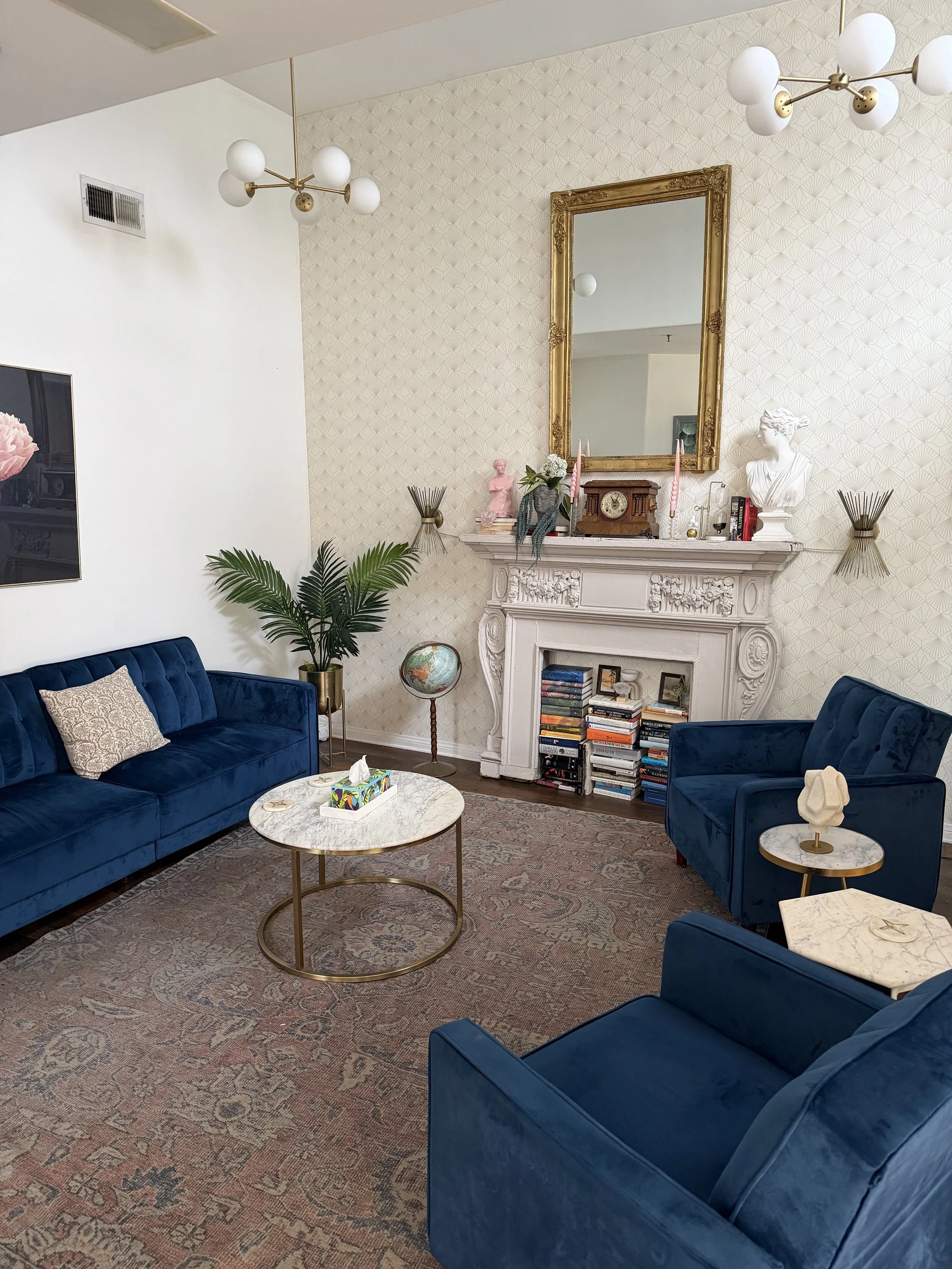 two dark blue velvet sofas, a white marble coffee table, a side table with decorative object, potted plant, ornate white fireplace mantle with a mirror, sculptures, books, and a globe, floral patterned rug, wallpaper with geometric pattern, chandeliers, and framed mirror.