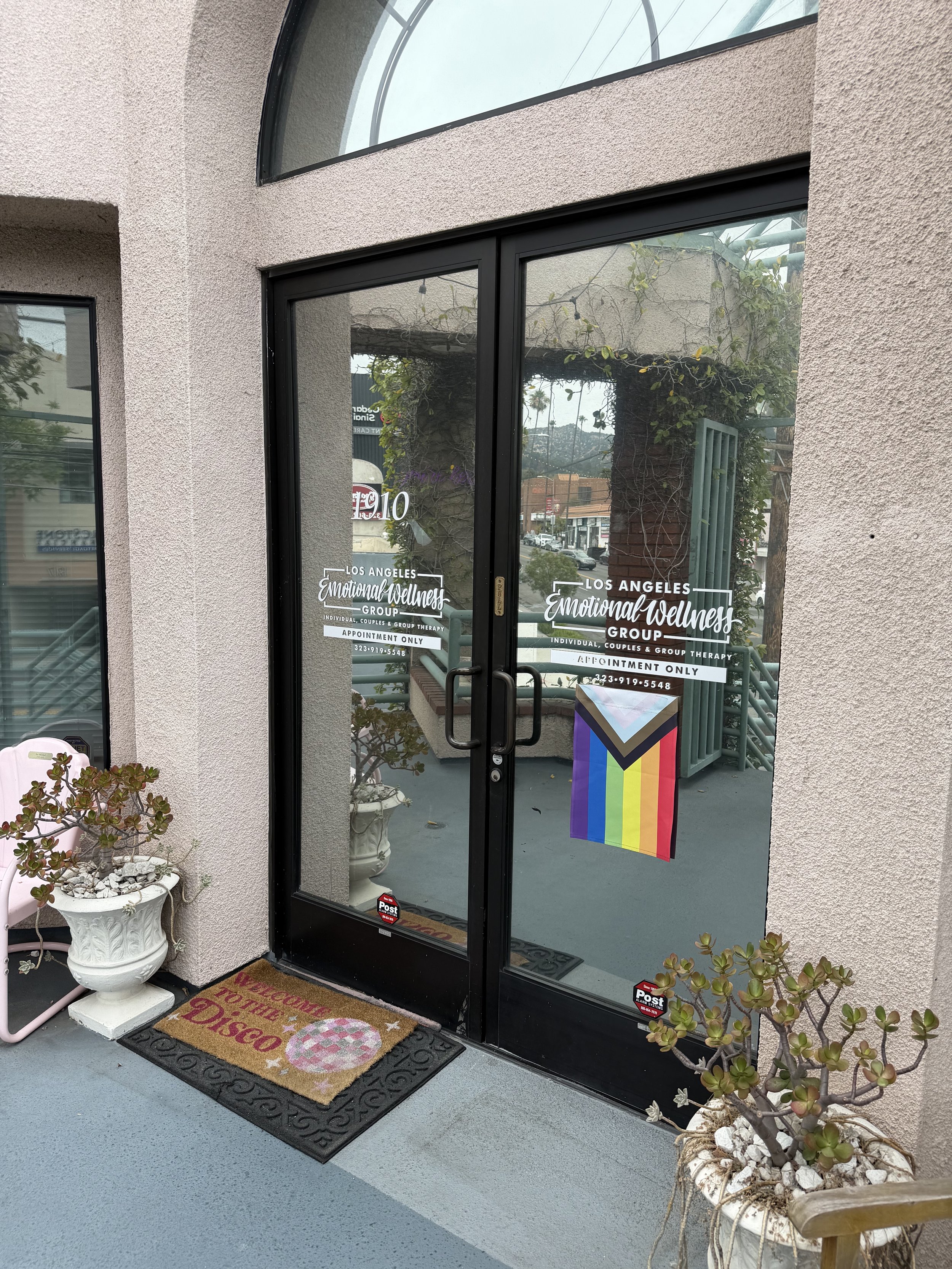 Black framed glass door with decals for Los Angeles Emotional Wellness Group, featuring a rainbow pride flag hanging on the right side of the door. Outside, there are potted plants and a welcome mat reading "Welcome to the Disco" on a light gray floor.