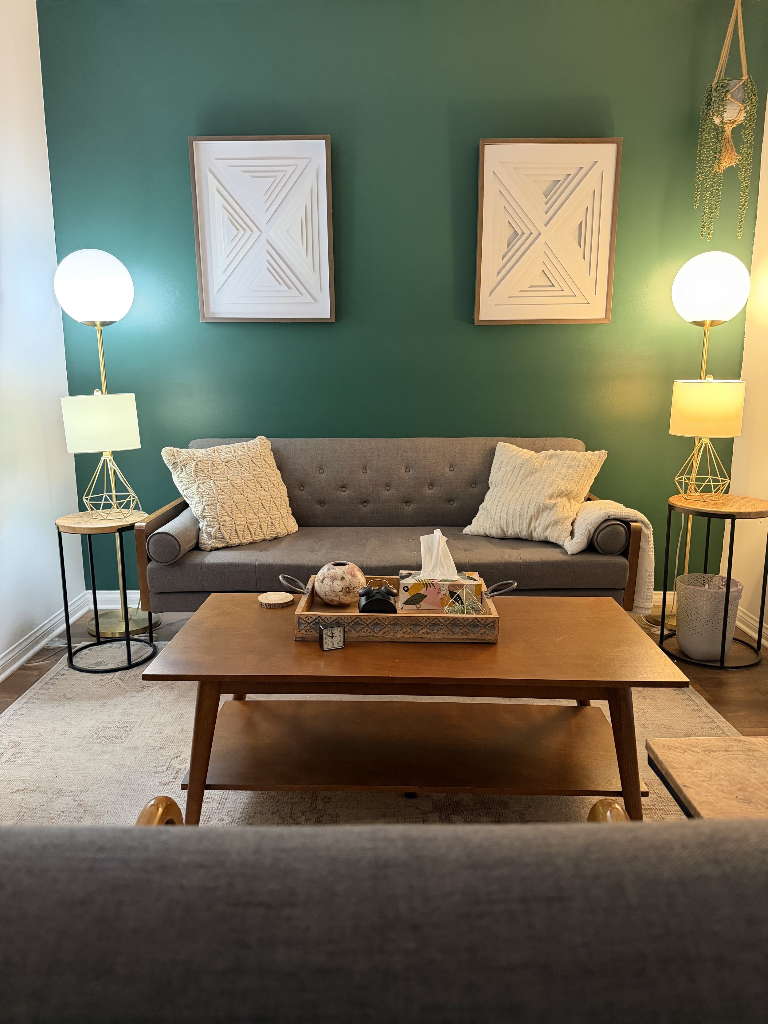Therapy office with green accent wall, two framed geometric art pieces, beige sofa with pillows, side tables with lamps, wooden coffee table with decorative items, hanging plant.