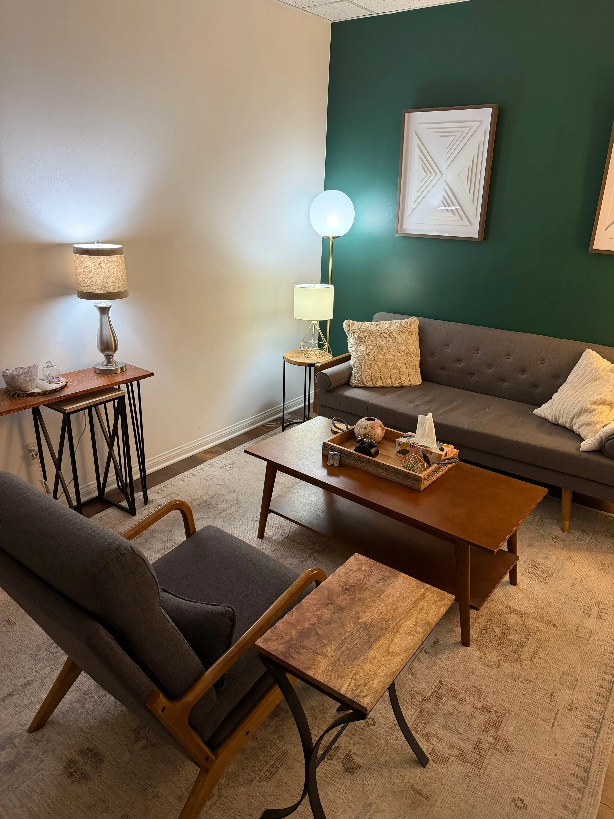 Therapy office with two sofas, one dark gray and one gray with pillows, wooden coffee table with decorative items, side table with a lamp, green accent wall with framed art, beige wall, and space heater.