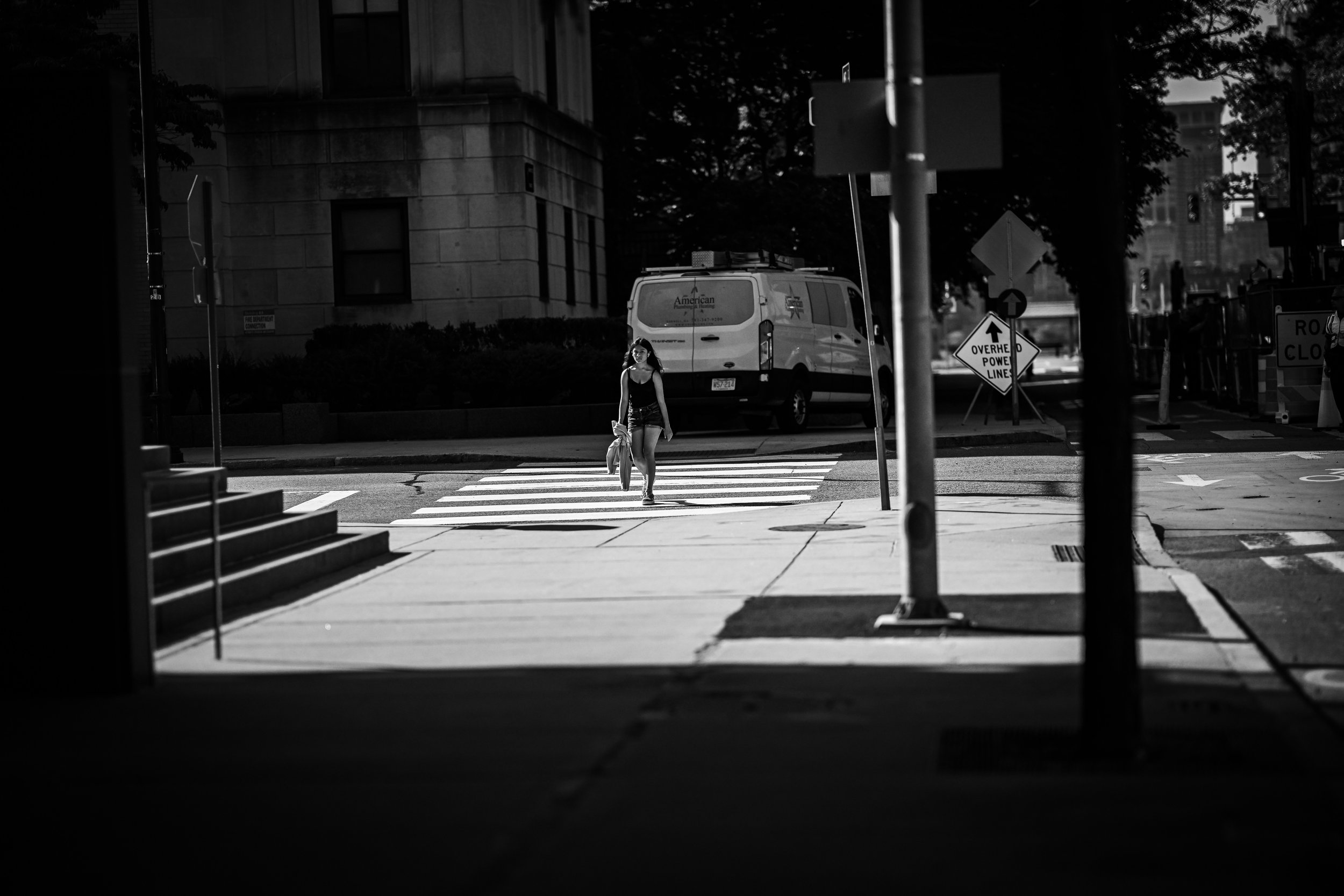A pedestrian crosses a sunlit city intersection framed by deep foreground shadow.