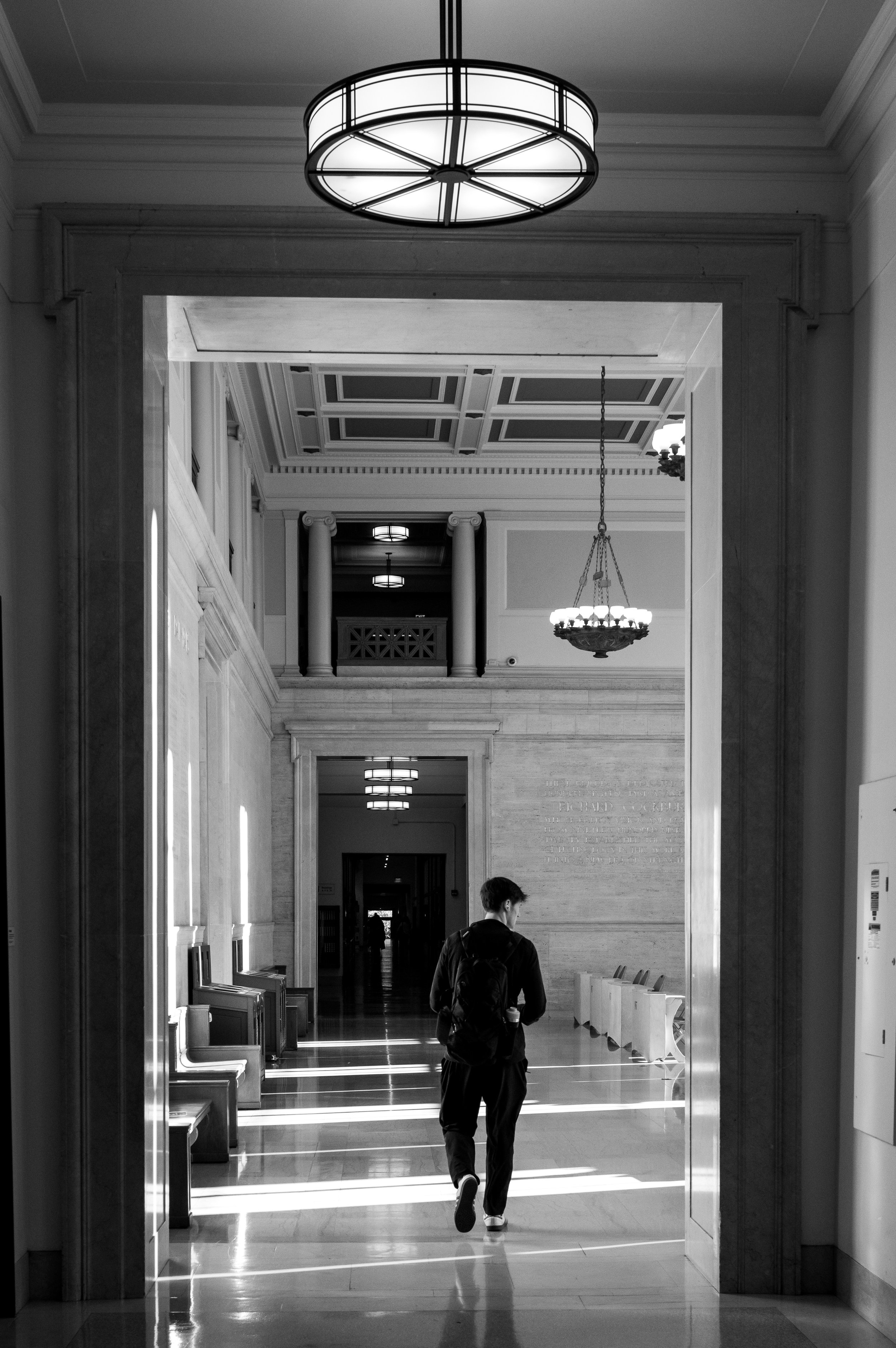 A lone person walks through a long, sunlit hallway beneath circular ceiling lights.