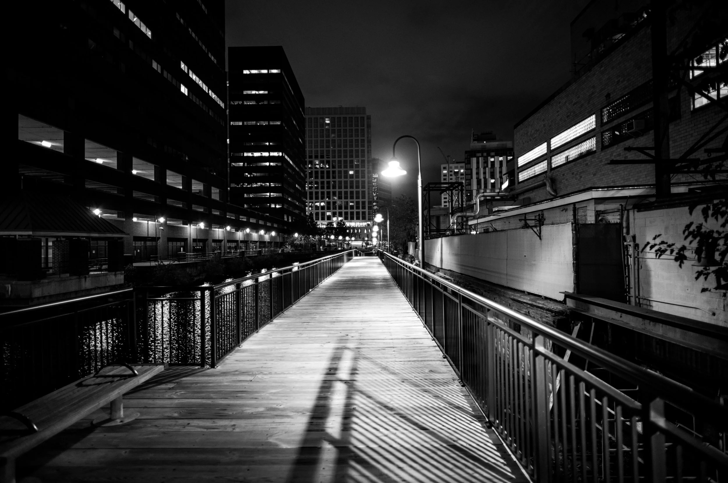 A brightly lit boardwalk and railing lead along a city canal at night.