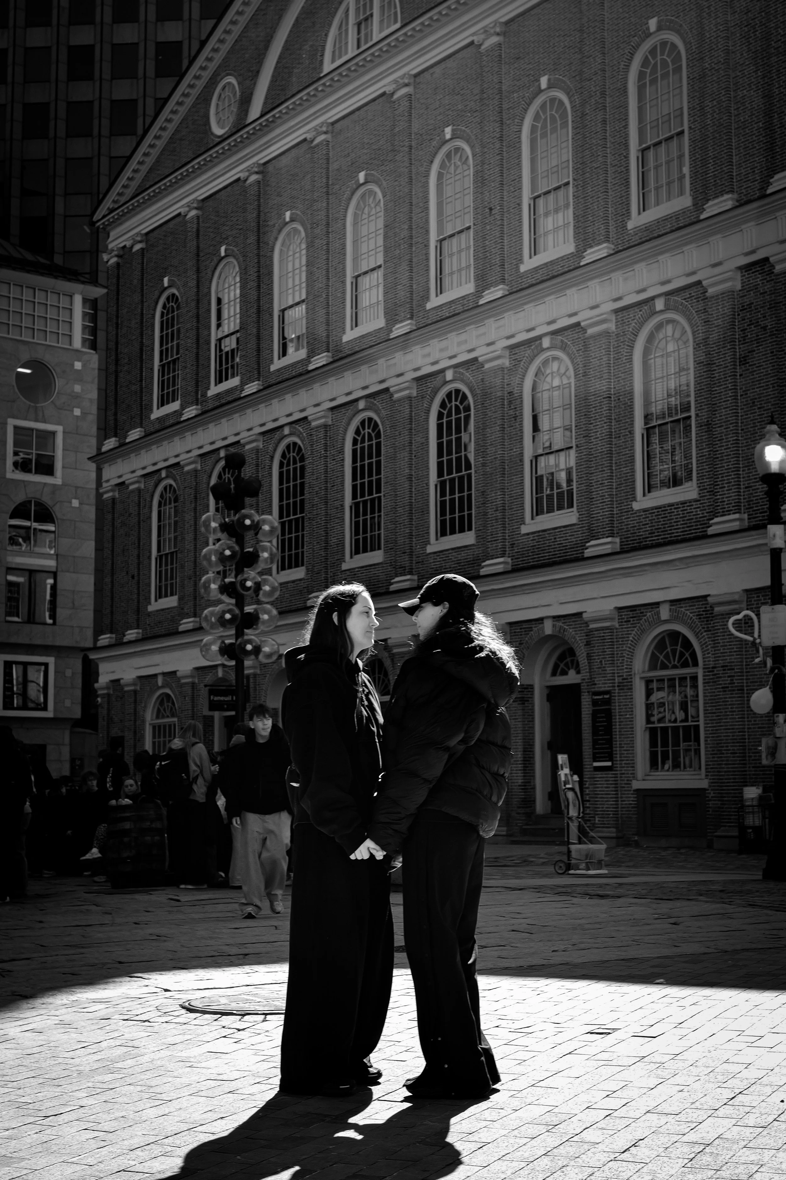 Two people stand together in sunlight before a historic brick building.