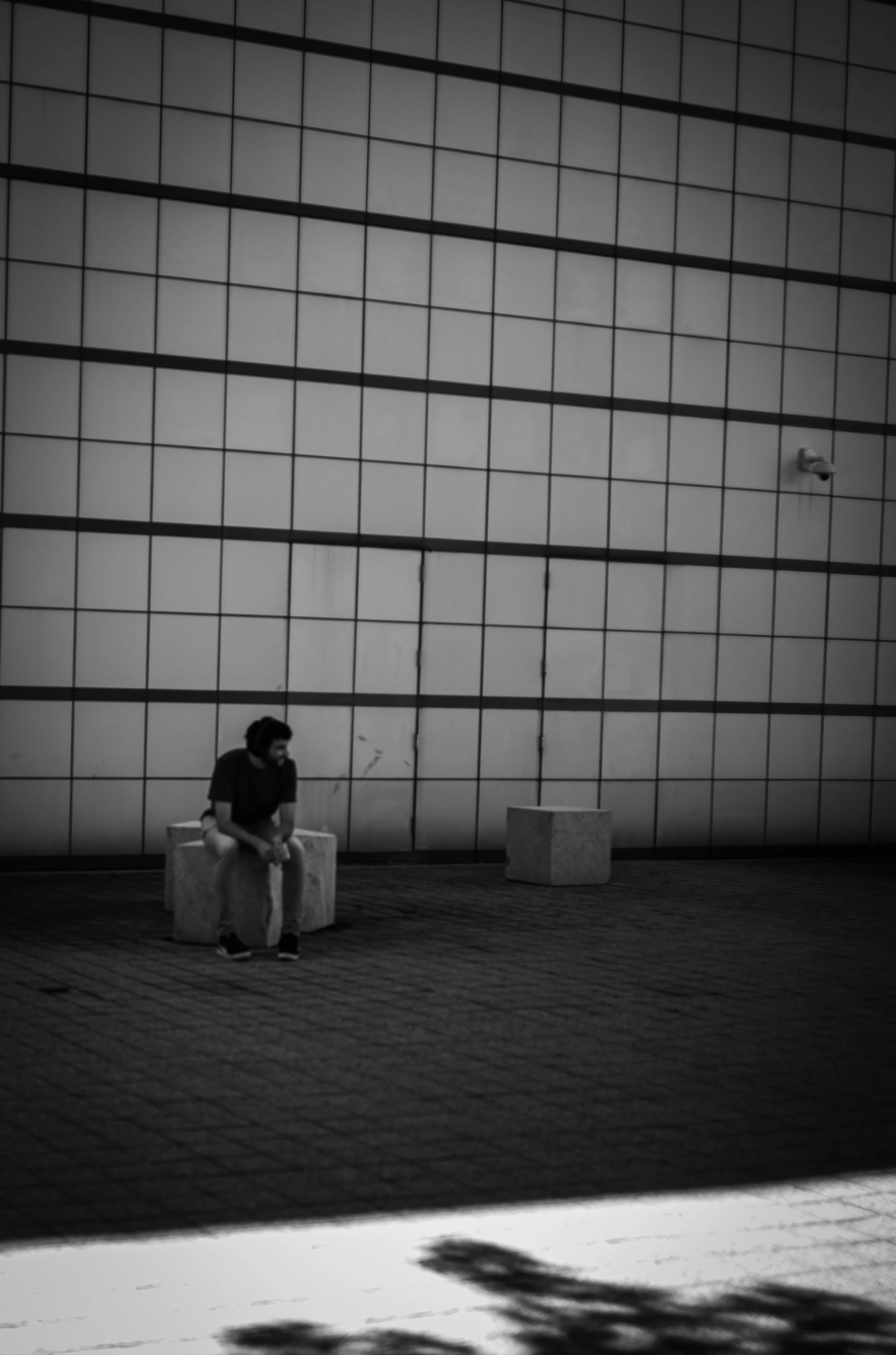A solitary person sits against a large grid-patterned wall in deep shadow.