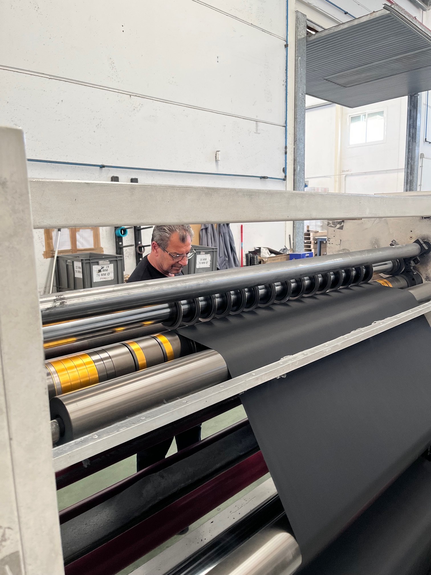 A man operating industrial machinery with large rollers in a factory or workshop environment, with white walls, a window, and various tools and equipment in the background.