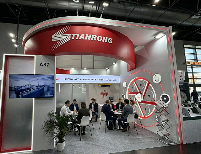 Trade show booth for Tianrong at an exhibition, featuring a curved red and white structure with company logo, a seating area with people in business attire, a wall display with monitors, a potted plant, and a stand with brochures or flyers.