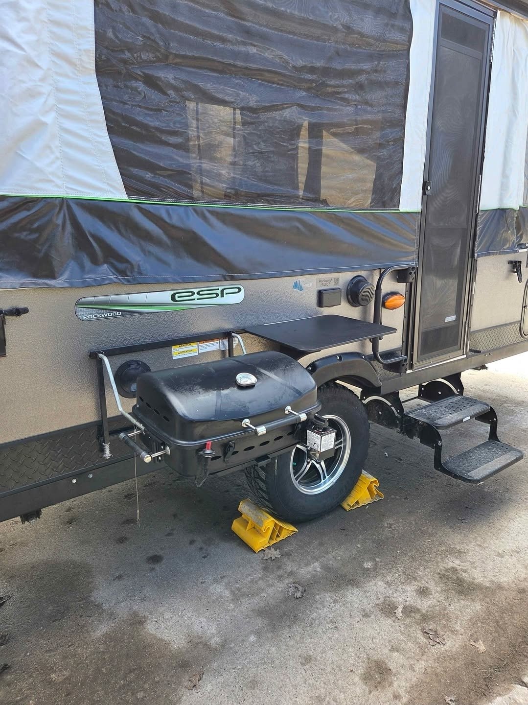 Close-up of the side of a travel trailer with a black barbecue grill mounted below the window, supported by yellow wheel chocks, and metal steps leading to a door.
