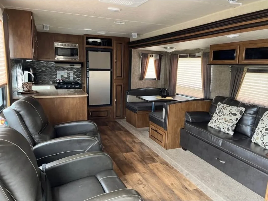 Interior of a recreational vehicle with leather chairs, wooden cabinets, a small kitchen area, and a dining area with bench seating and a table.