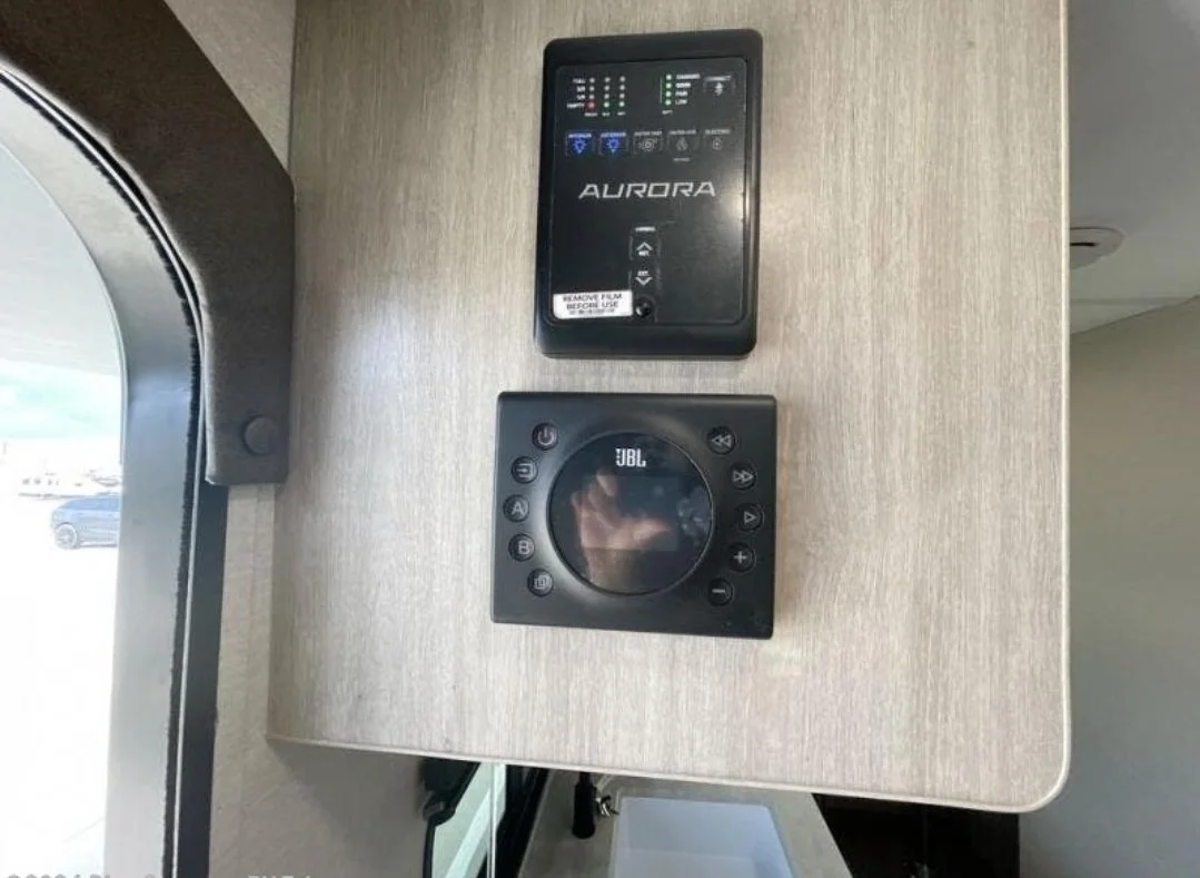 A control panel and a JBL speaker mounted on a light wood-colored wall next to a window within an RV or camper.
