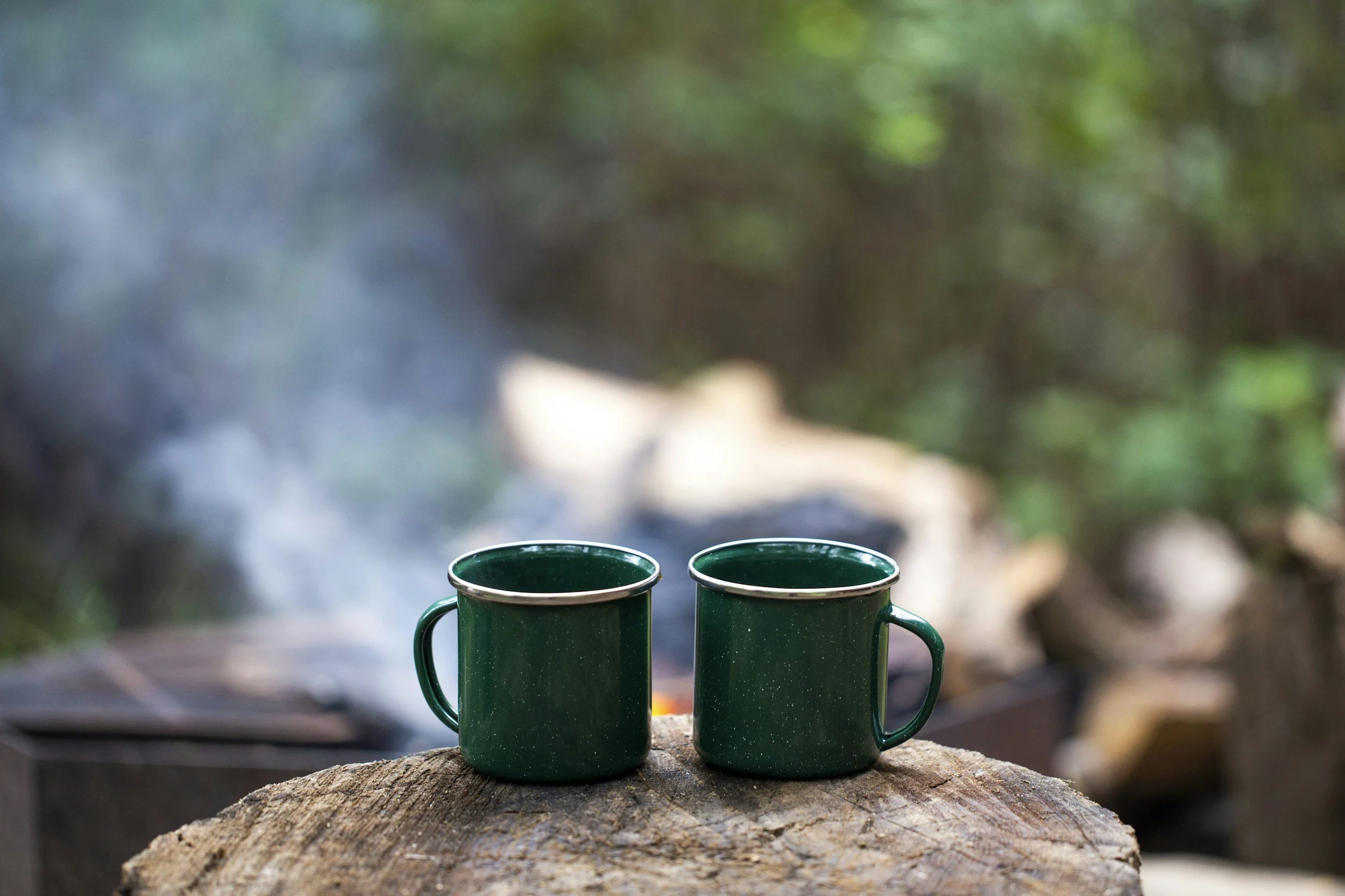 Two green enamel coffee mugs placed side by side on a rough wooden surface outdoors, with a blurred background of trees and foliage.
