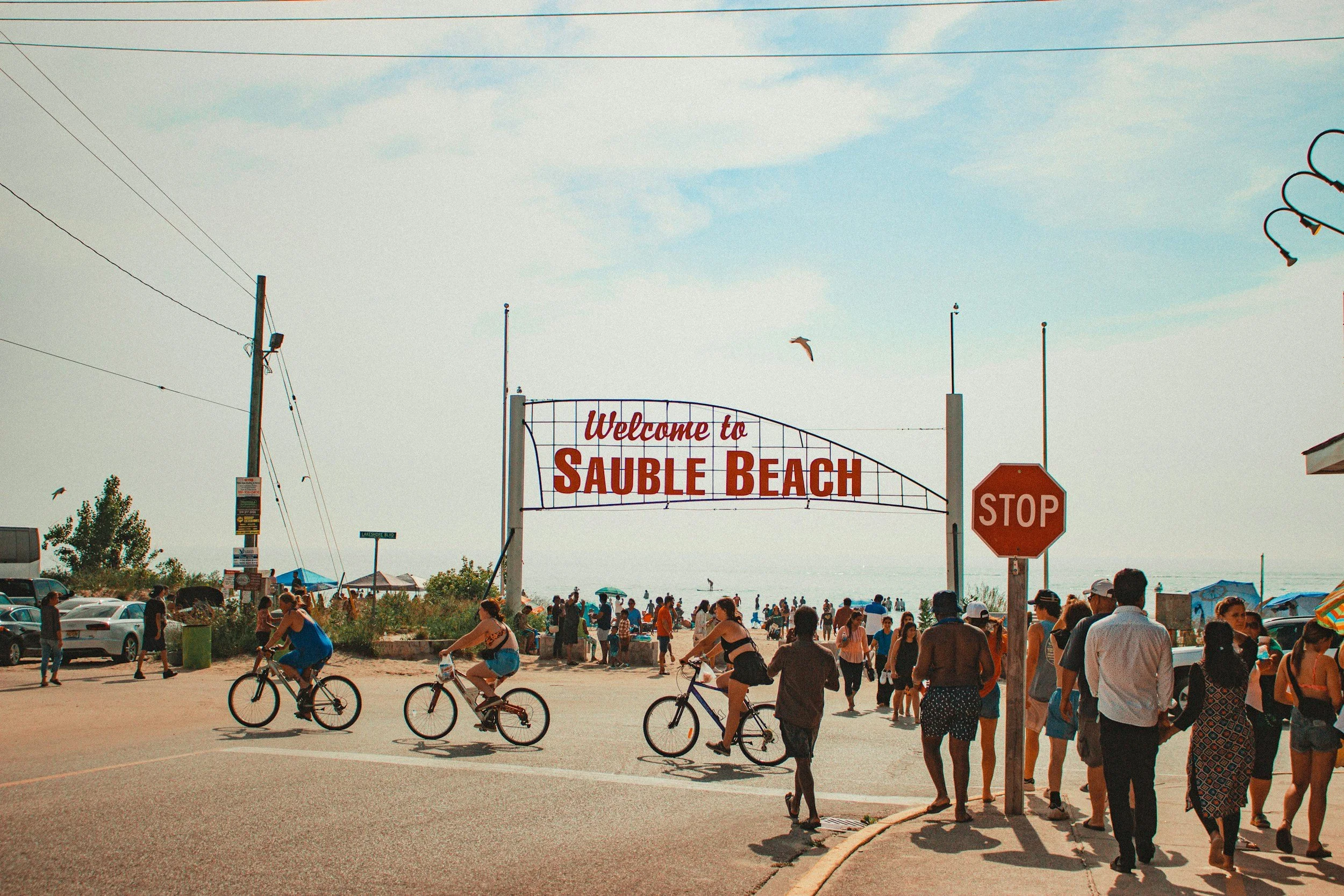 Crowd of people at Sauble Beach, including children riding bicycles, in front of the sea with some tents and umbrellas, a large sign reads 'Welcome to Sauble Beach,' overcast sky, stop sign, and utility poles.