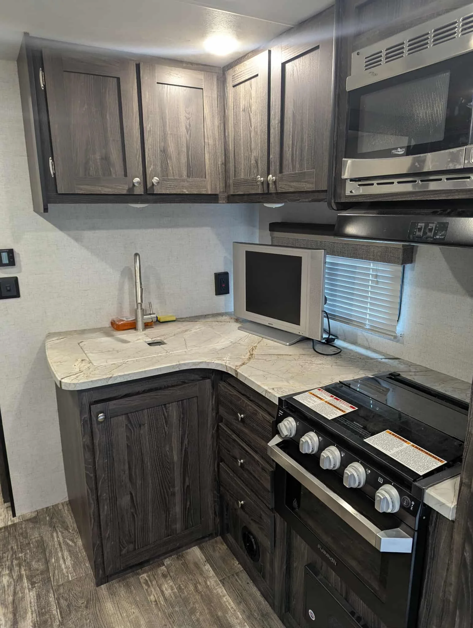 A kitchen with dark wood cabinets, a marble countertop, a small TV, and a stainless steel oven with a stovetop. There is a window with blinds and a sink with a faucet.