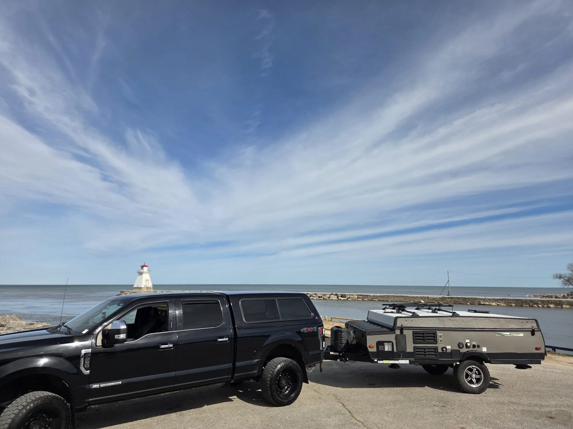 A scenic view of a lighthouse at the bay with a blue sky and wispy clouds. A black pickup truck with a trailer attached is parked in the foreground near the shore.
