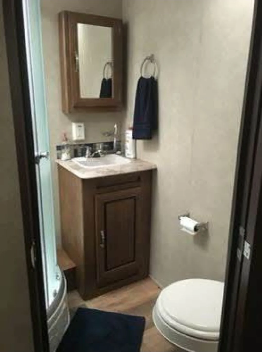 A small bathroom with a wooden vanity, marble countertop, and mirror cabinet above the sink. Towel hanging on a ring, toilet paper holder with paper, a toilet, and a dark bathmat on the wooden floor.