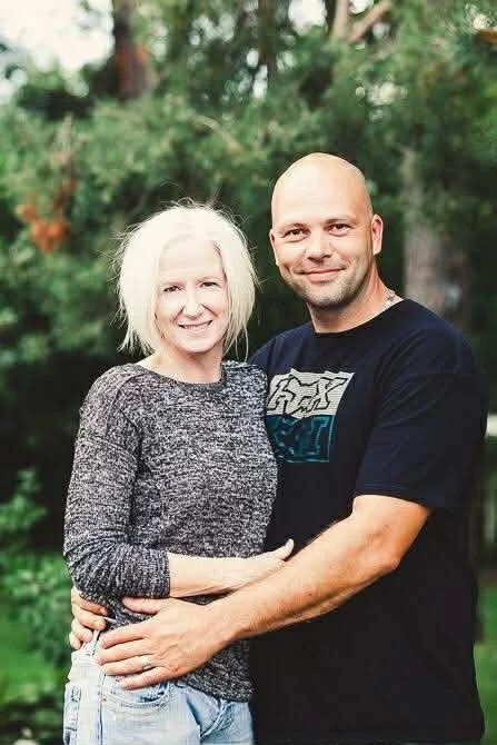 A woman and a man embracing outdoors in front of trees, smiling at the camera.