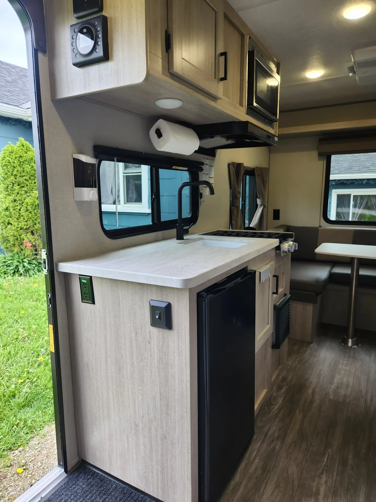Interior view of a small RV kitchen with a countertop, sink, coffee maker, microwave, and cabinets. Outside view of green grass and a house with a blue exterior through the open door.