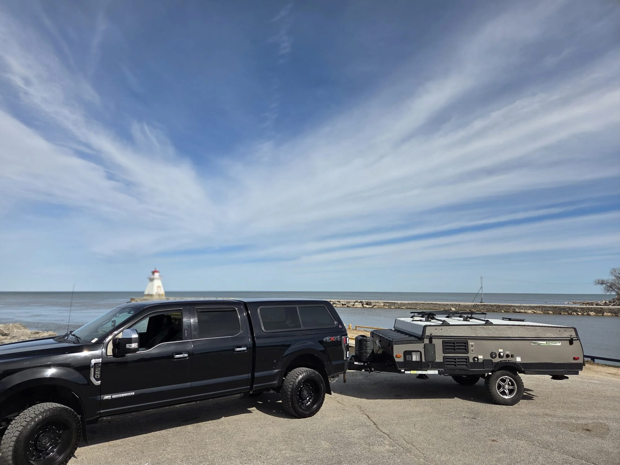 A black truck parked by a waterfront with a trailer attached, a lighthouse in the background, and the sky with scattered clouds.