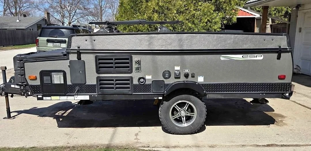A gray and black portable camping trailer parked on a driveway with a vehicle and trees in the background.