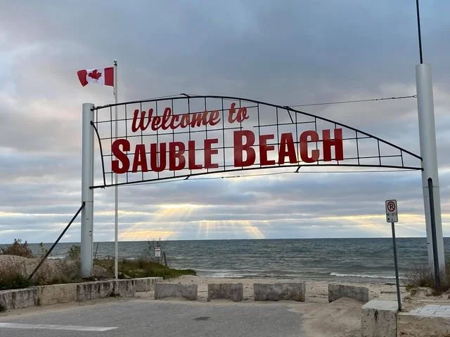 Entrance sign reading 'Welcome to Sauble Beach' with a Canadian flag, overlooking the water with a cloudy sky.