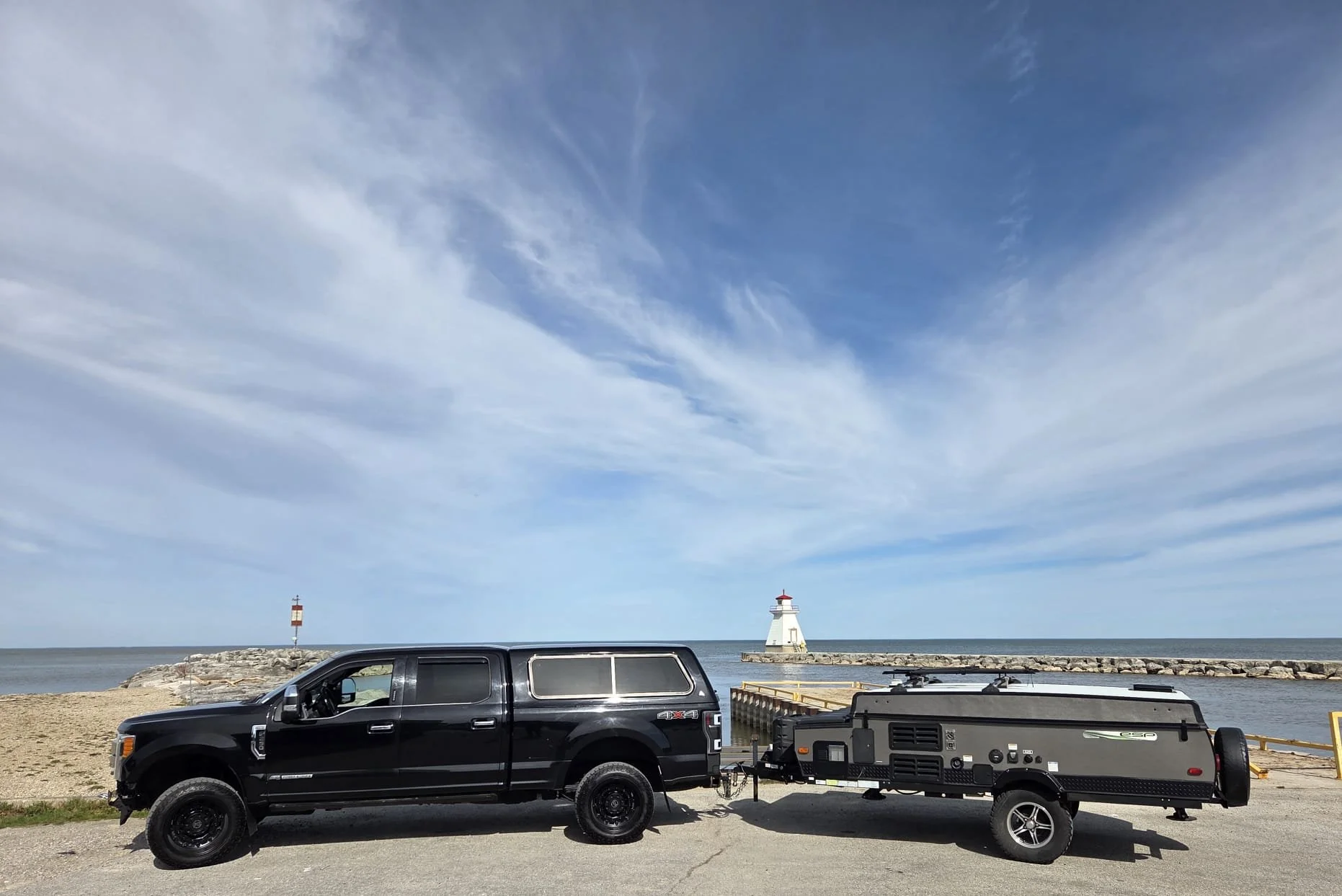 A black pickup truck towing a trailer parked near a beach with a lighthouse and a smaller lighthouse in the background under a partly cloudy sky.