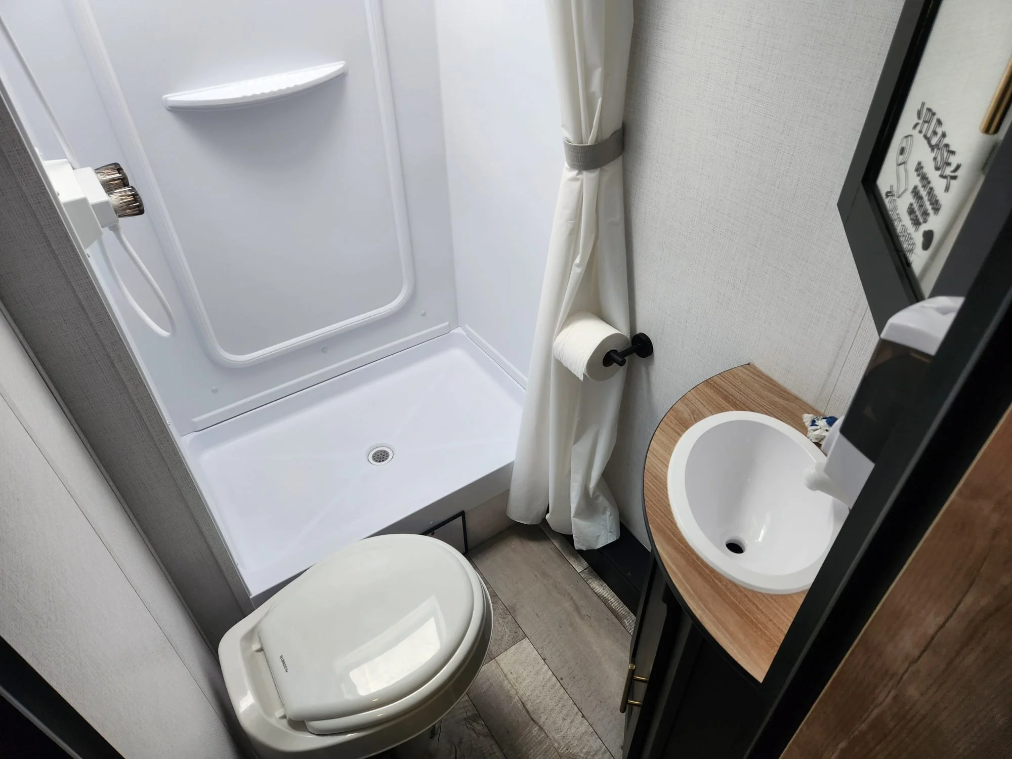 A compact bathroom with a shower stall, a toilet, a small sink with a wooden countertop, a toilet paper holder, a foldable shower curtain, and a mirror on the wall. The space is efficiently organized in a recreational vehicle or small apartment.