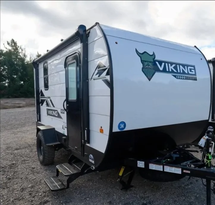 A white Viking travel trailer with black accents, parked outdoors on gravel with a step ladder and hitch in the foreground.
