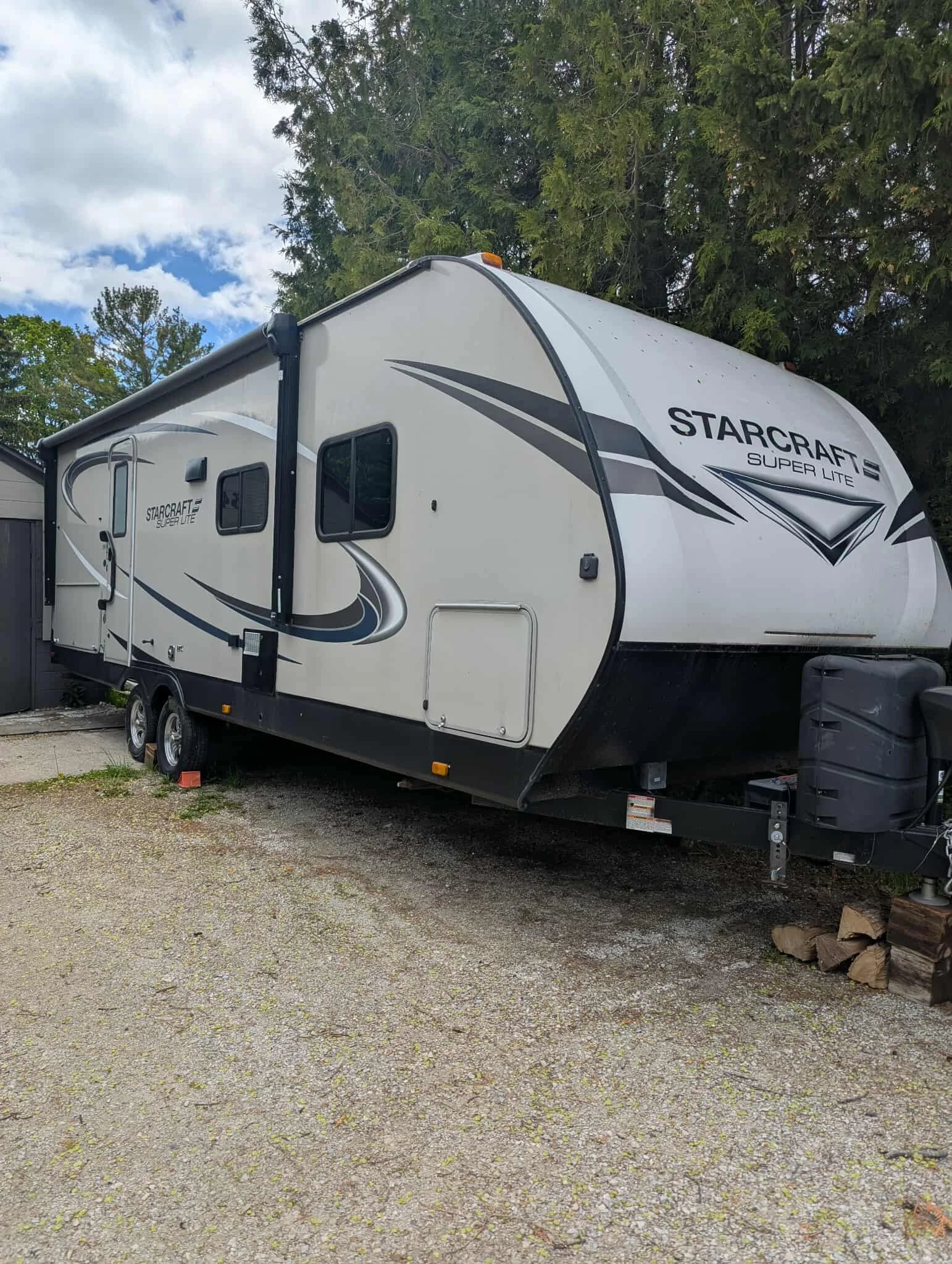 A white Starcraft Super Lite travel trailer parked outdoors on a gravel surface, with trees and a partly cloudy sky in the background.
