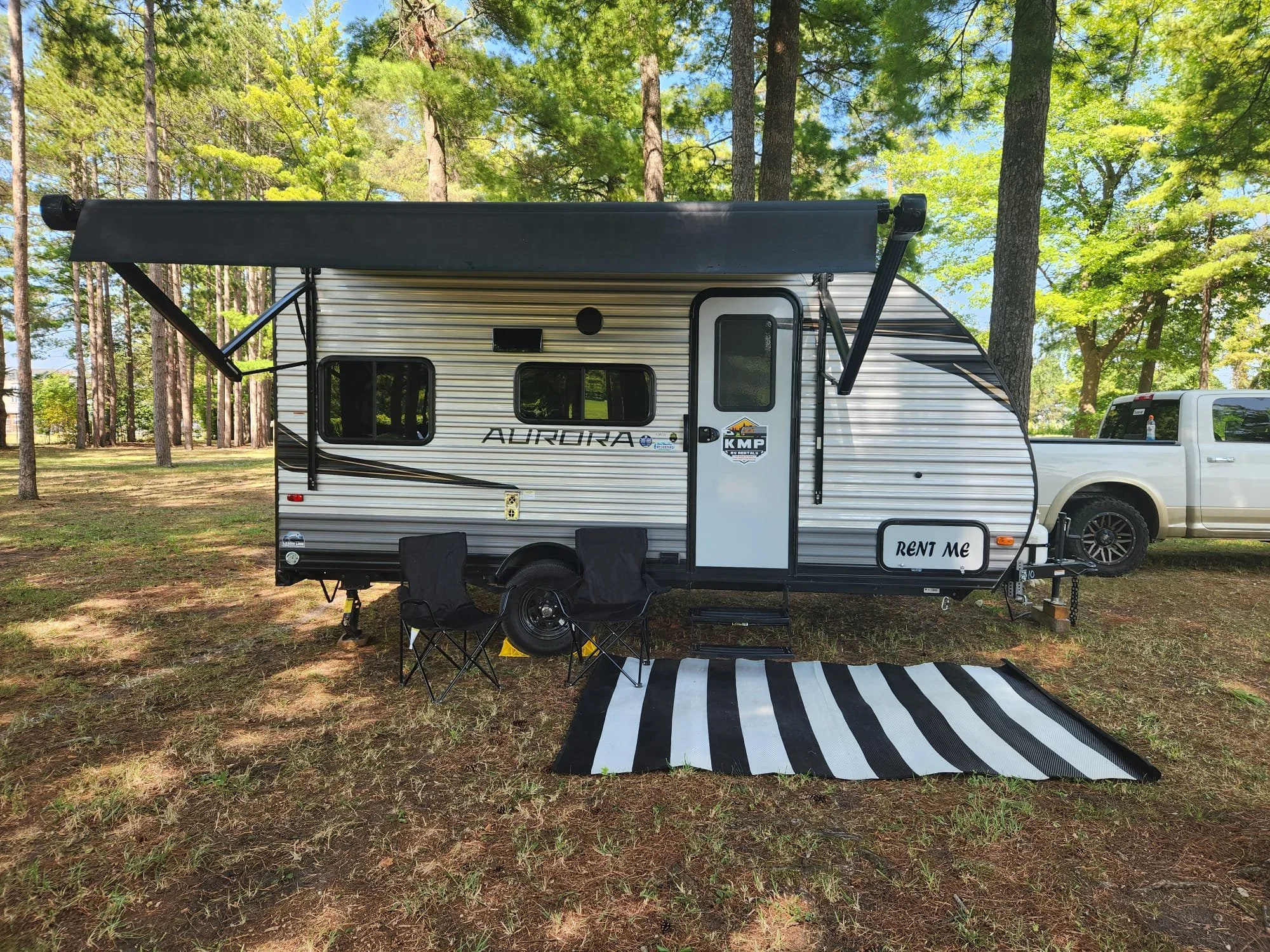 A small travel trailer named 'Aurora' is parked in a wooded area with tall trees and green foliage, under a clear blue sky. There are two black chairs, a striped outdoor rug, and a sign that says 'Rent Me' in front of the trailer.